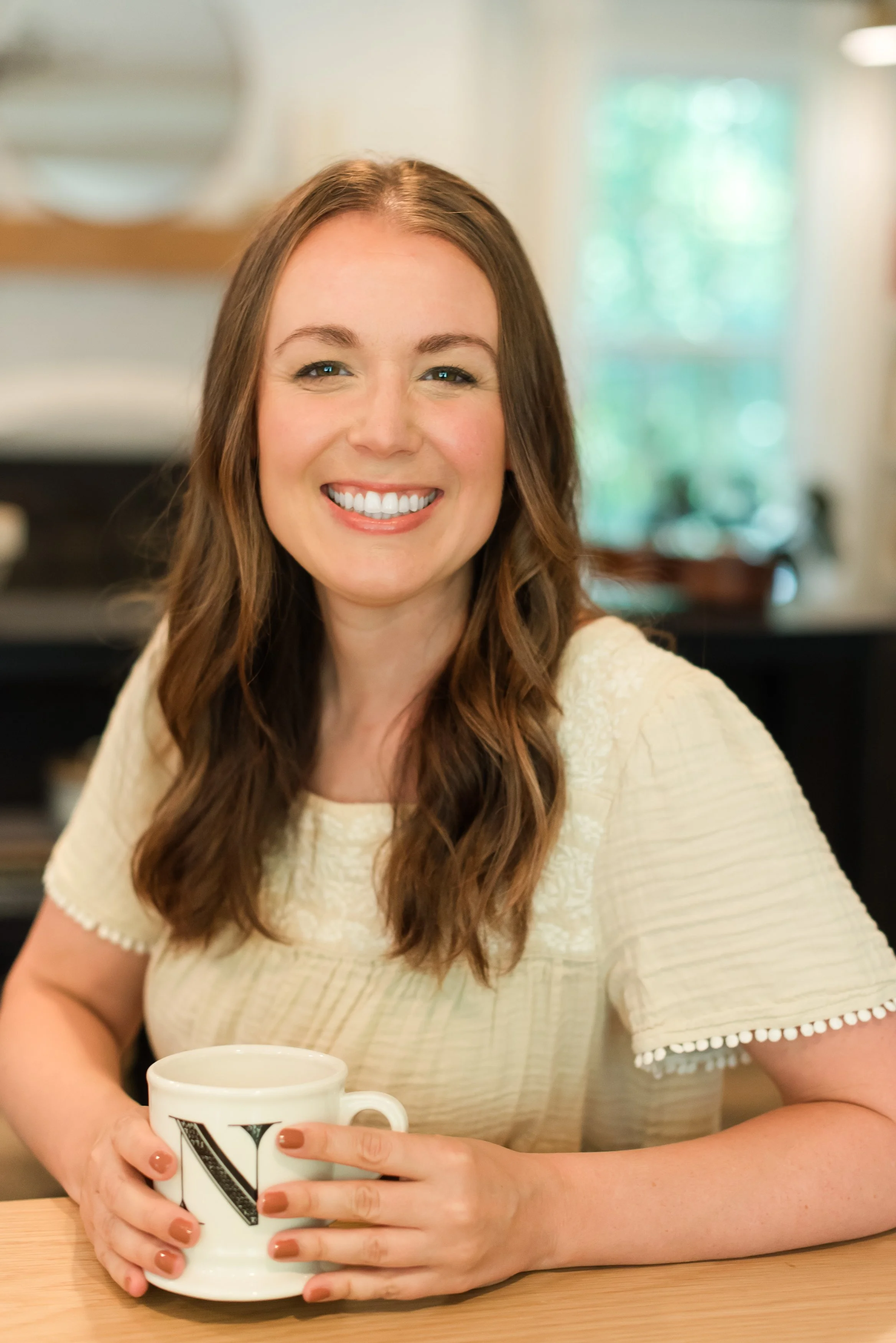 A woman with long wavy brown hair smiling, sitting at a table holding a white mug with a letter 'V' on it, in a cozy, well-lit room.