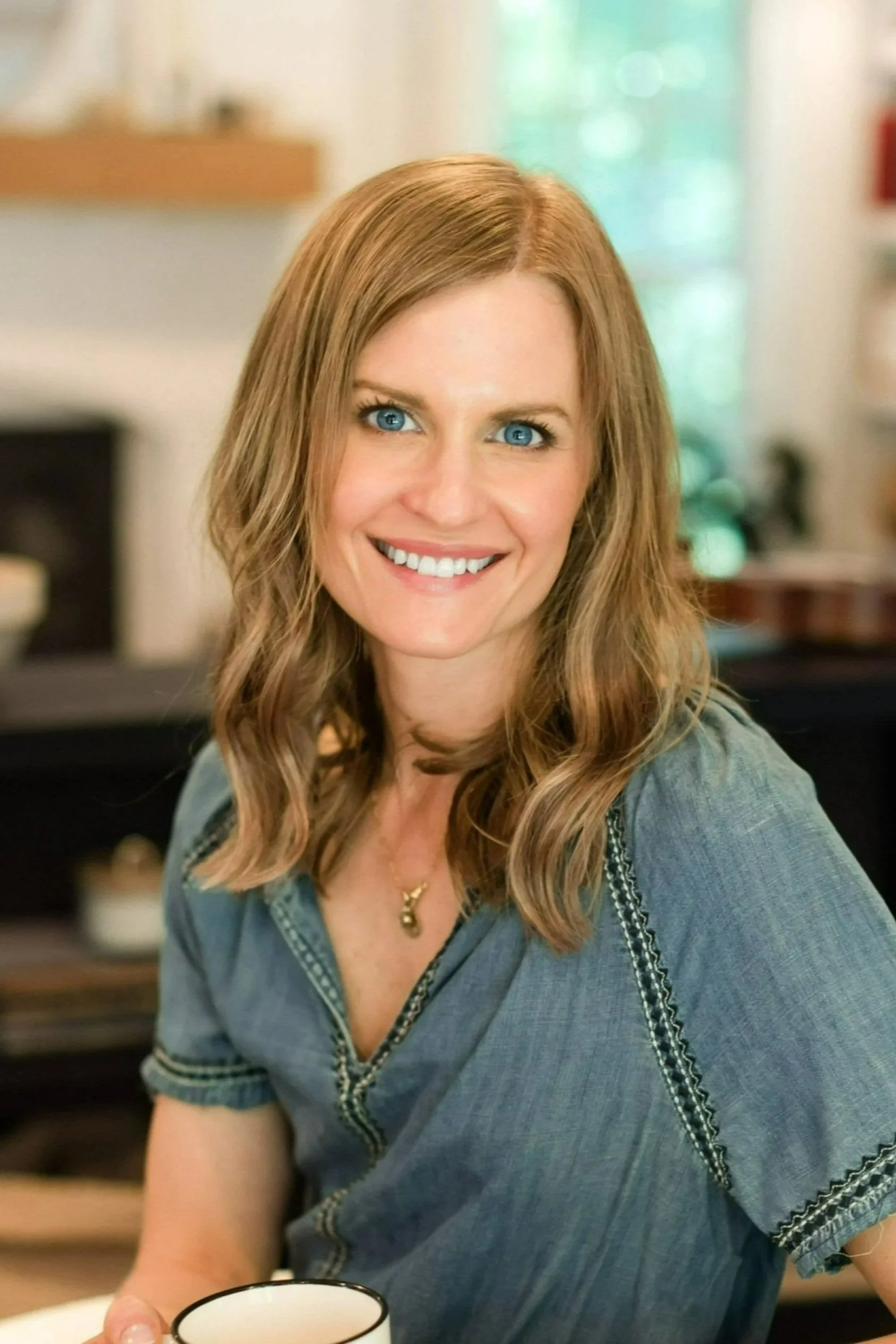 A woman with long, wavy red hair and blue eyes smiling and holding a cup, sitting in a cozy room with natural light, blurred background.