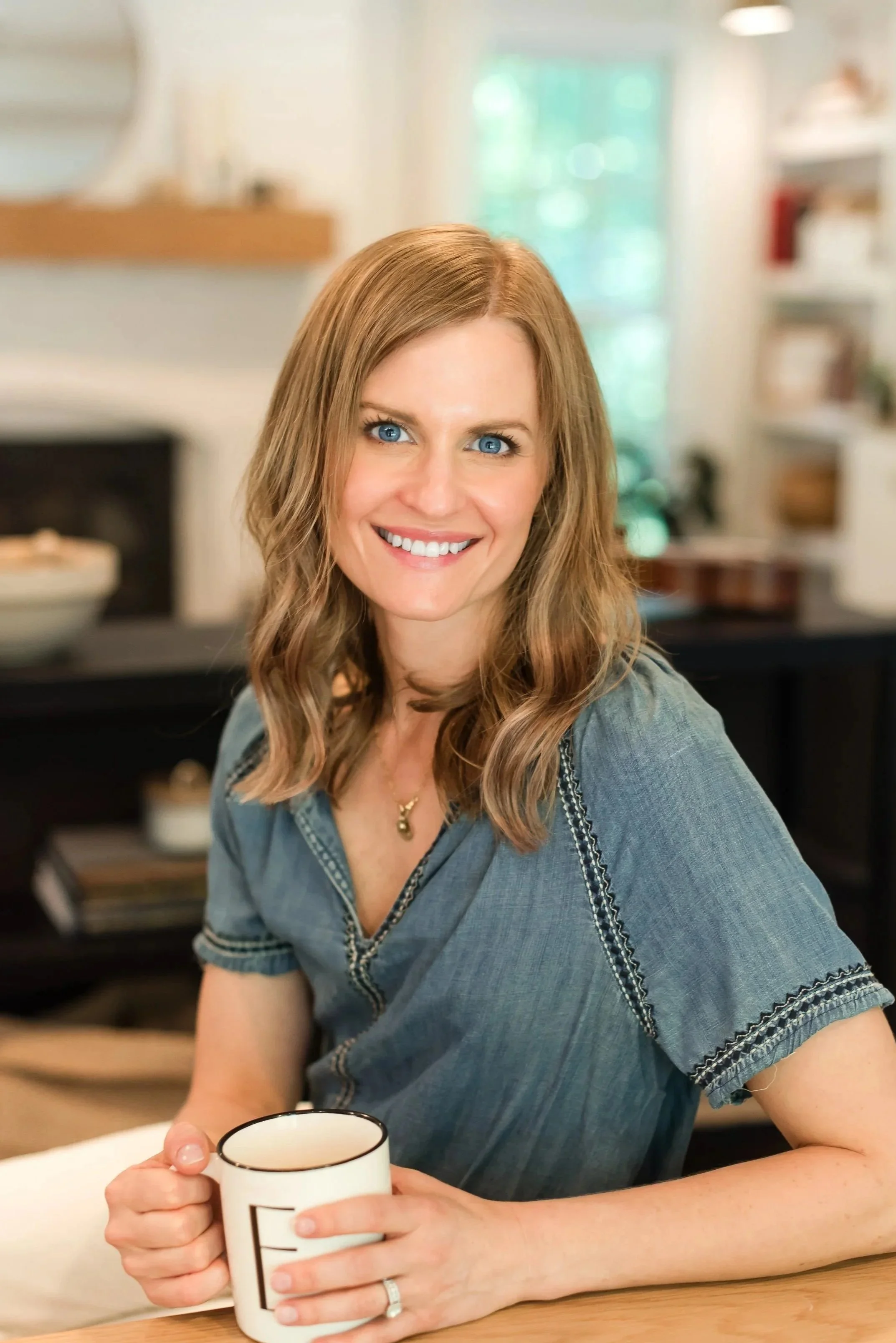A woman with shoulder-length wavy brown hair, blue eyes, and a bright smile sits at a table holding a white mug with black accents. She is wearing a blue denim shirt with decorative stitching and a gold necklace. The background shows a cozy, well-lit kitchen or living space with shelves and a door with glass panes.
