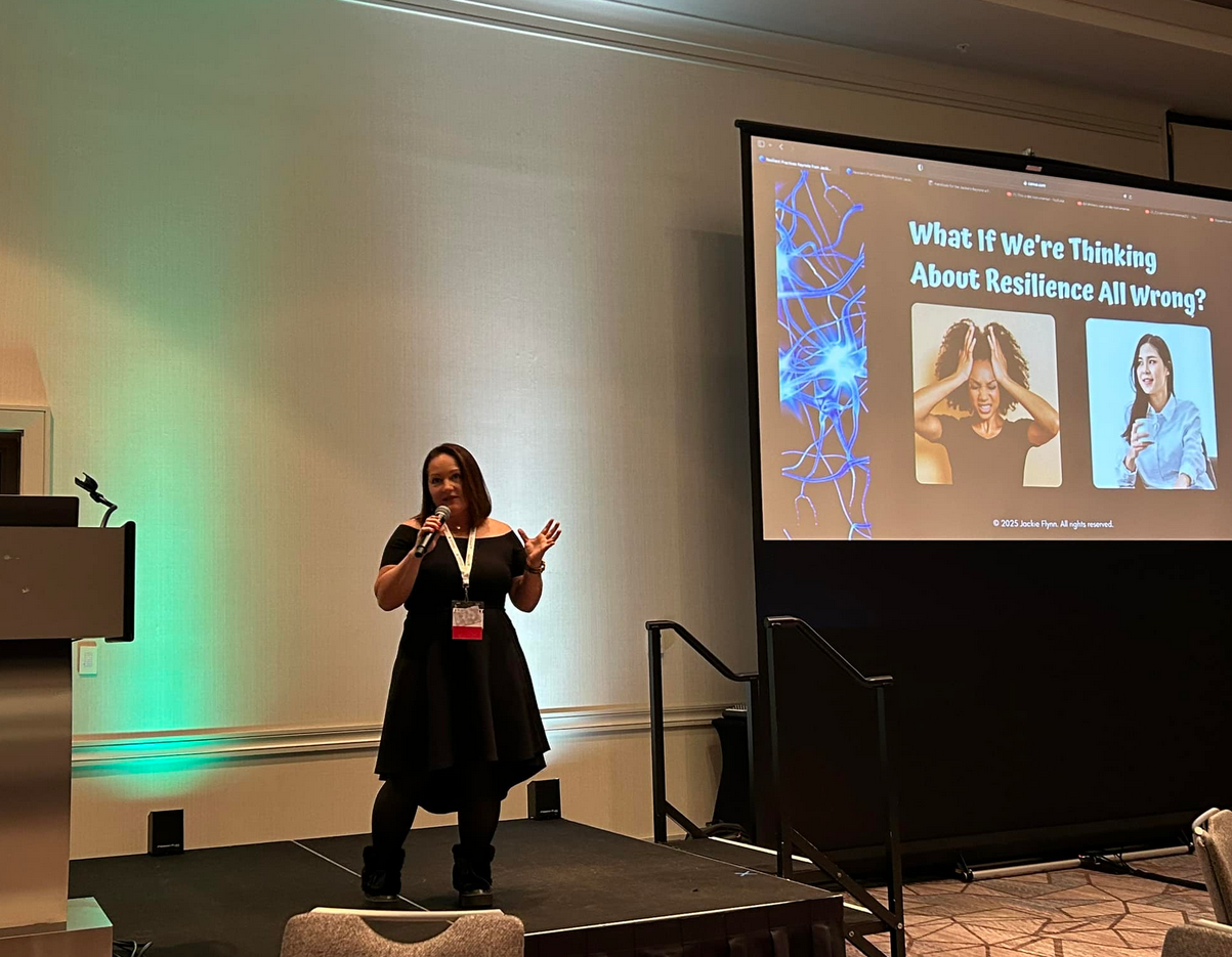 A woman standing on a stage holding a microphone, giving a presentation in a conference room. A large screen behind her displays a slide titled 'What if We're Thinking About Resilience All Wrong?' with images of a frustrated woman and a smiling woman.
