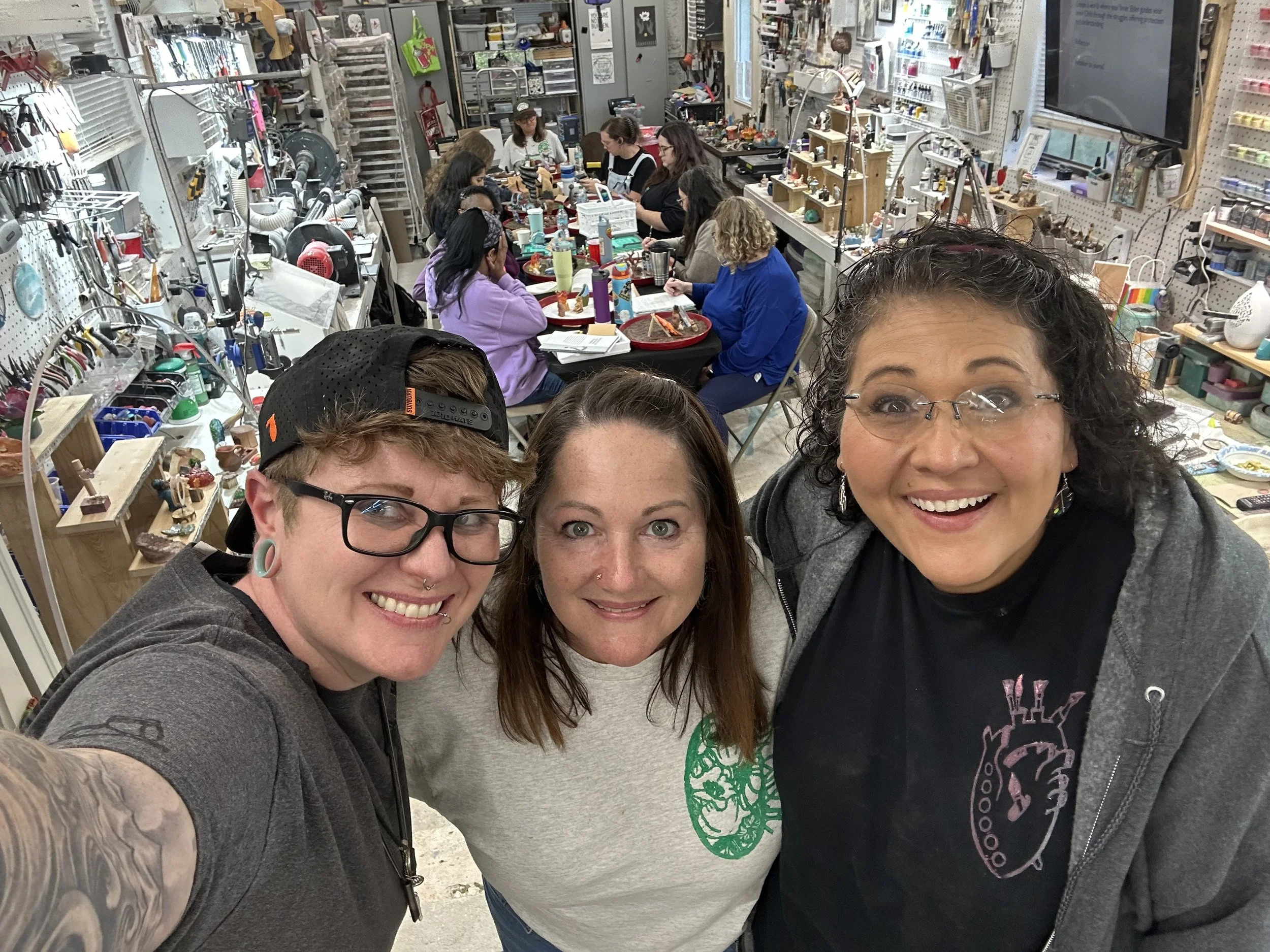 Three women taking a selfie in a craft workshop, with women working on projects at a large table in the background.