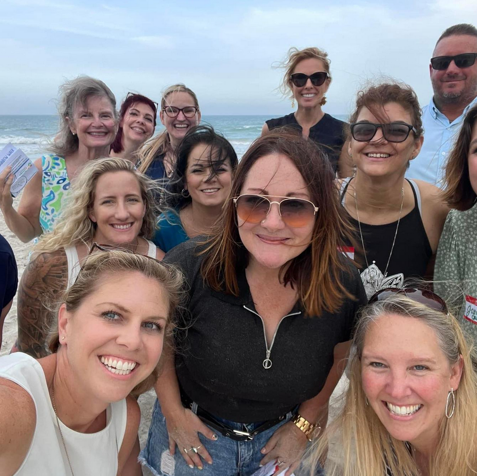Group of women and one man smiling at the beach, dressed casually, with the ocean in the background.