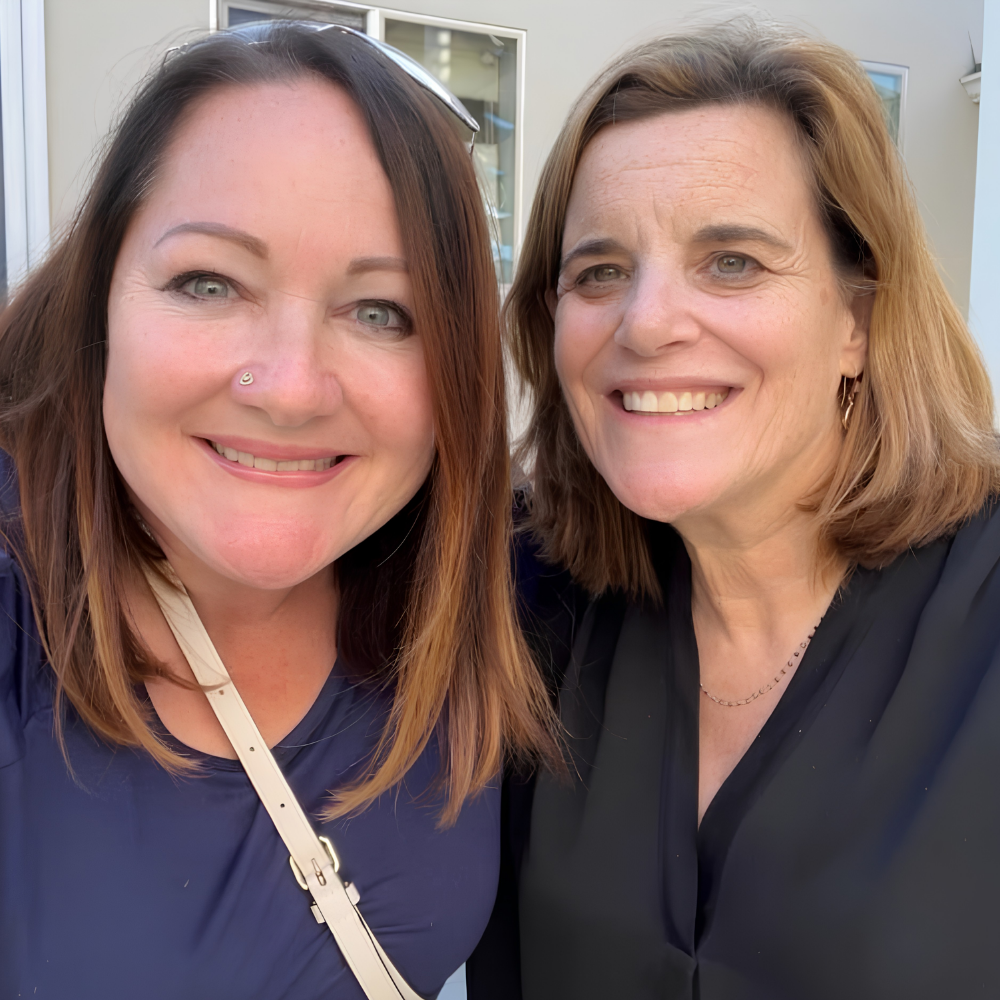 Two women smiling outdoors, close-up selfie with buildings in the background.