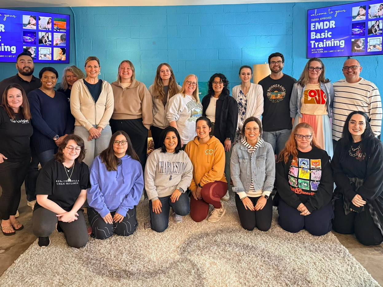 Group of 19 diverse women and men posing indoors in front of blue wall with screens displaying 'EMDR Basic Training'