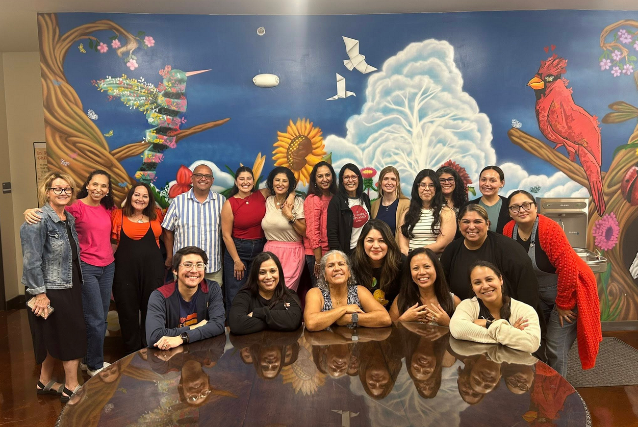 Group photo of 17 diverse people smiling and posing indoors in front of a colorful mural featuring a tree, birds, squirrels, a sunflower, and a pink flamingo, with table in front.