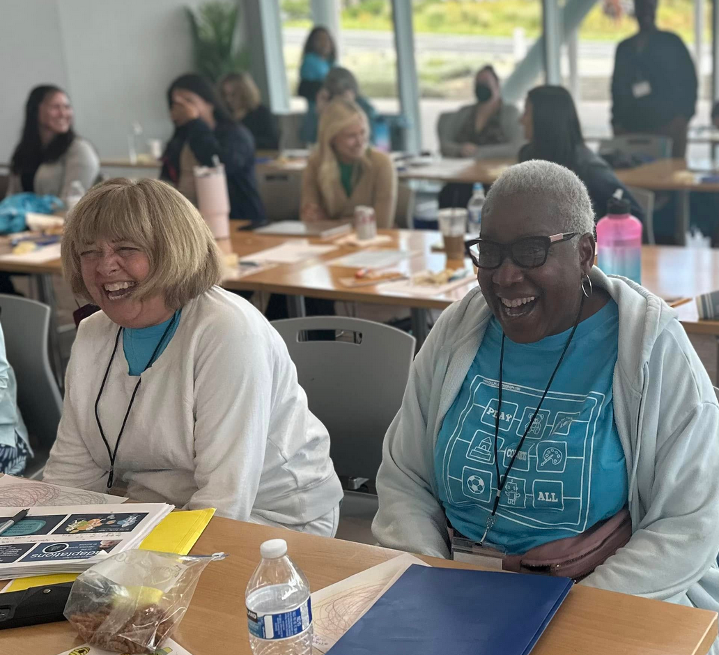 Two women laughing while sitting at a table in a bright, modern room with other people in the background. The woman on the left has blonde hair and is wearing a white jacket, and the woman on the right has gray hair, glasses, and is wearing a blue shirt and white hoodie.