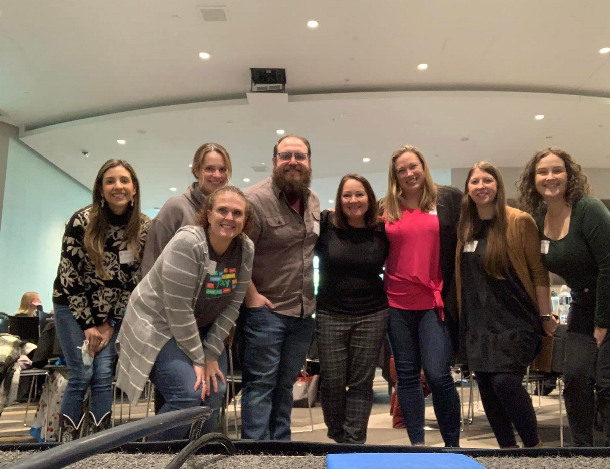 Group of eight smiling people, five women and one man with glasses and beard, standing indoors in a spacious, brightly lit room.