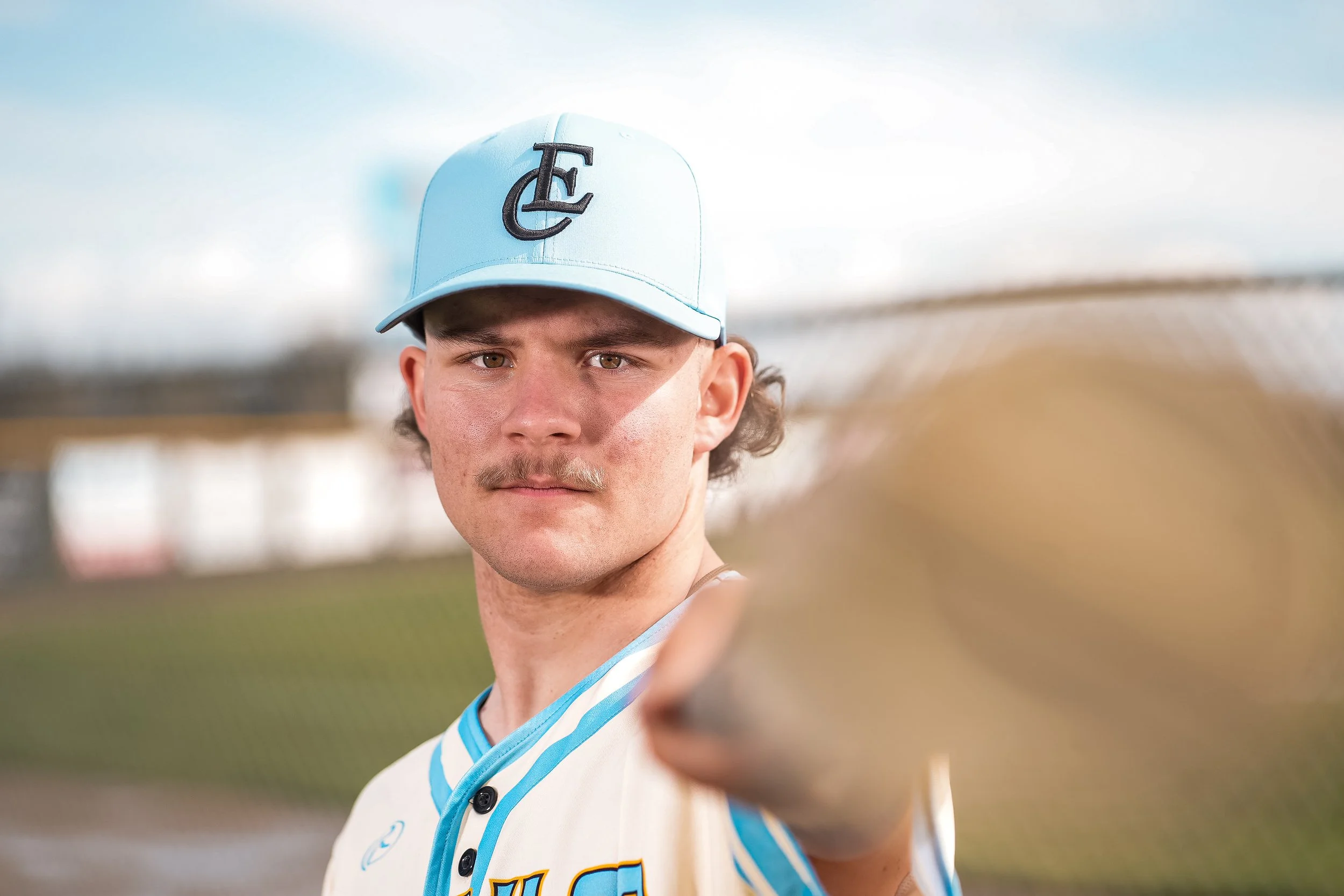 A young man with curly brown hair and a light mustache is wearing a white baseball jersey with blue accents and a white baseball cap with a black emblem, standing outdoors under a clear sky.