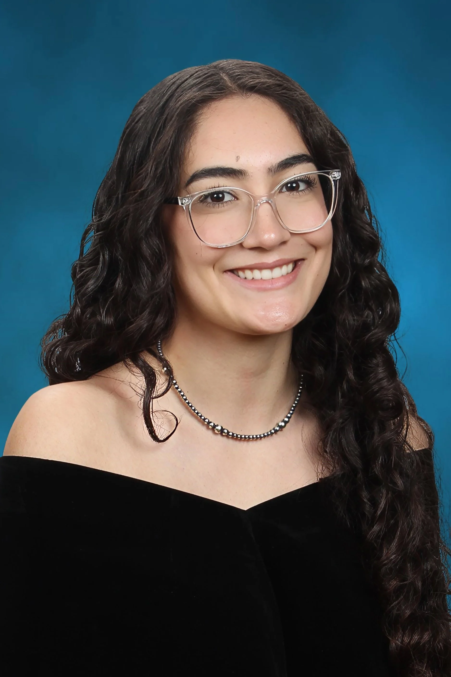 A young woman with long, curly dark hair wearing silver-rimmed glasses, a black off-the-shoulder top, and a beaded necklace, smiling against a blue background.
