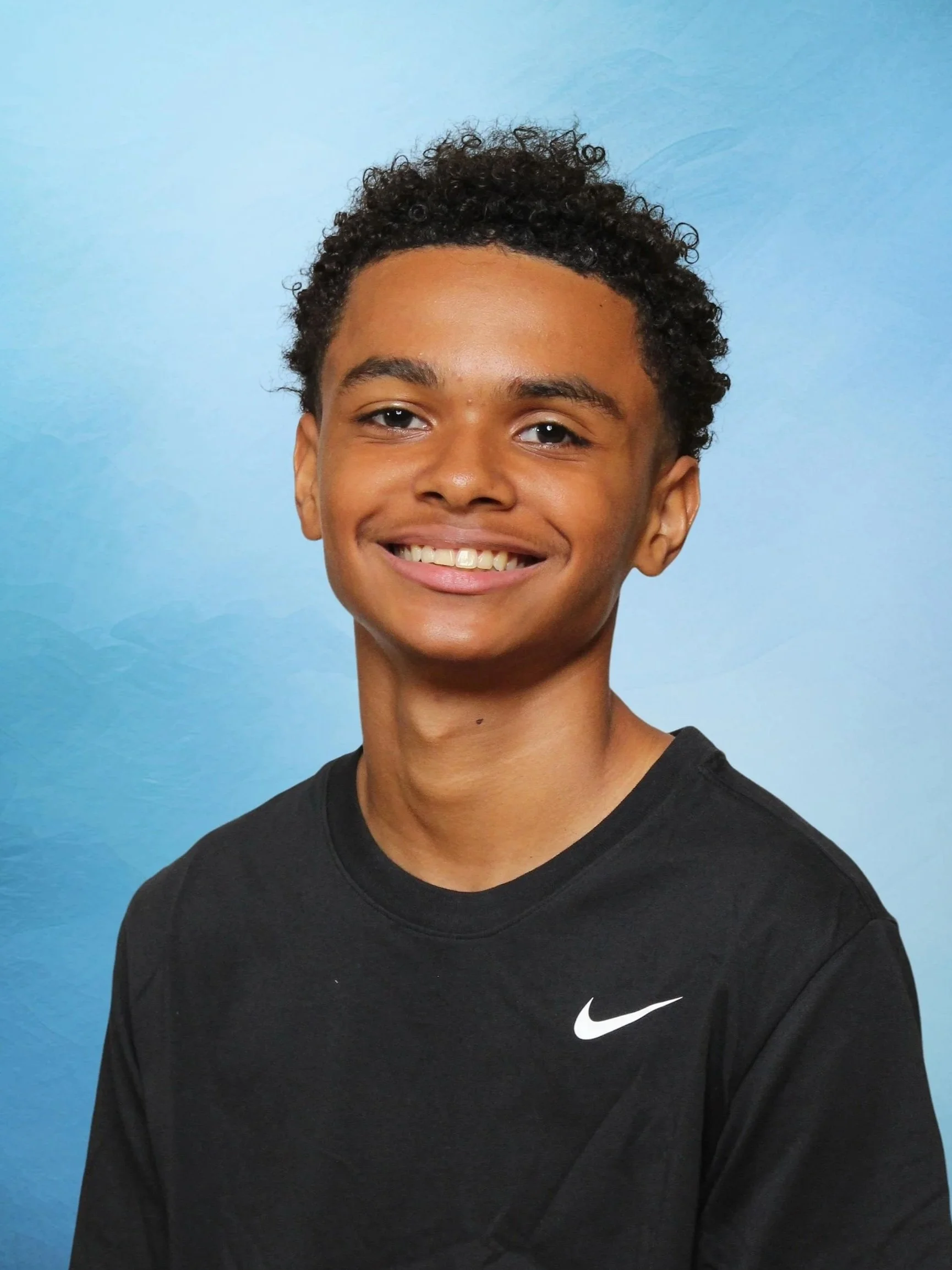 Close-up of a young person with short curly hair smiling, wearing a black Nike shirt, against a light blue background.