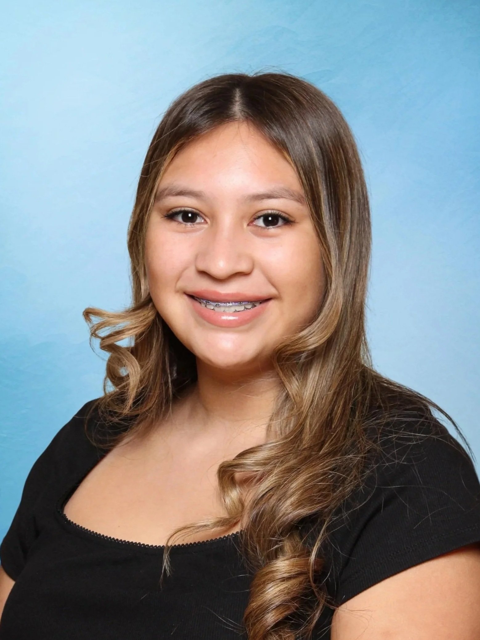 Close-up of a young woman with long, wavy hair, wearing a black top, smiling against a blue background.