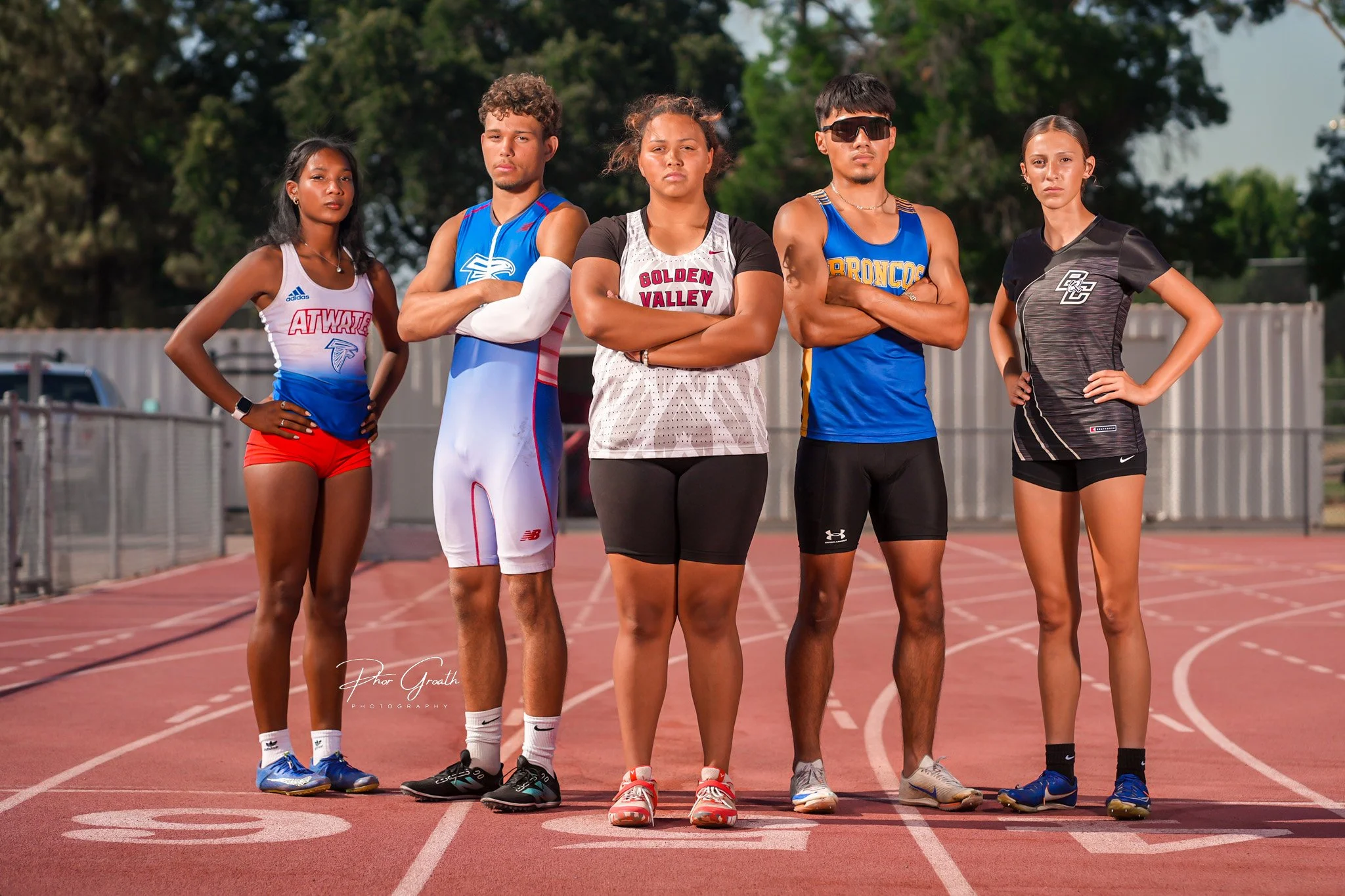 Five young athletes standing on a track, arms crossed, wearing athletic clothing, ready for a race with trees in the background.