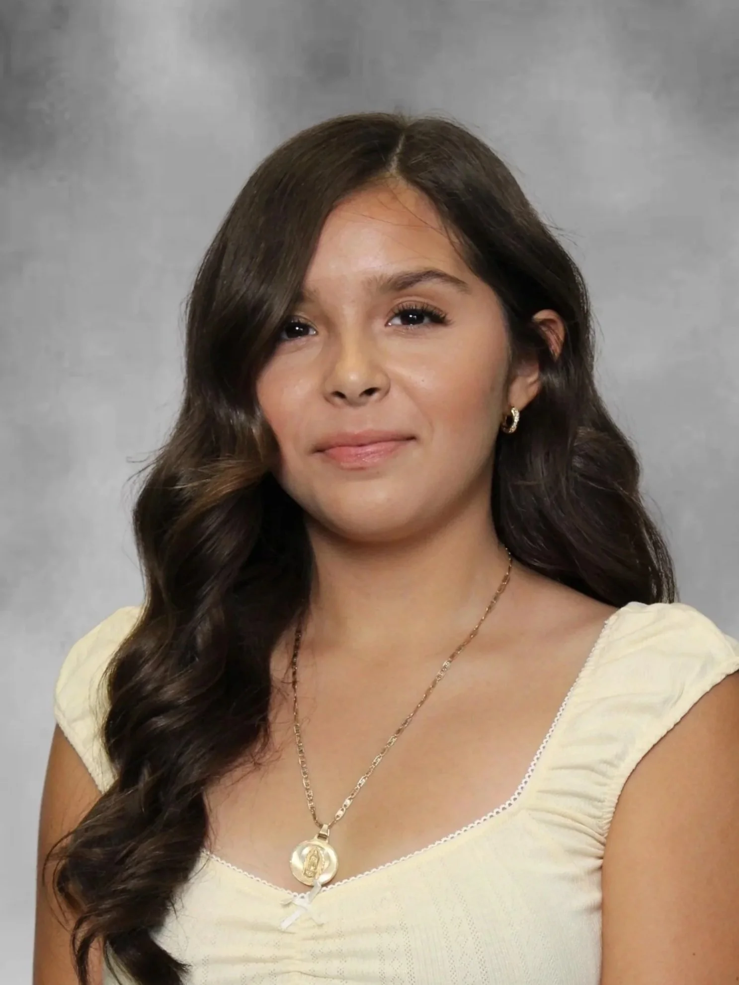 Close-up of a woman with long, dark, wavy hair, wearing a yellow top with white trim, gold earrings, a gold necklace, and a medallion pendant, against a gray background.