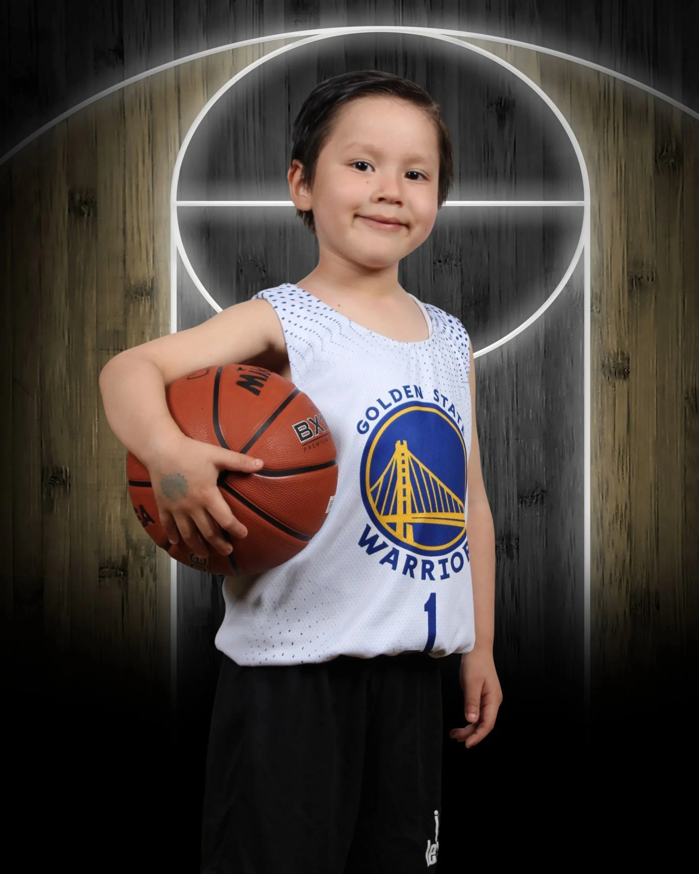 A young boy wearing a Golden State Warriors basketball jersey holding a basketball, standing in front of a basketball hoop backboard.