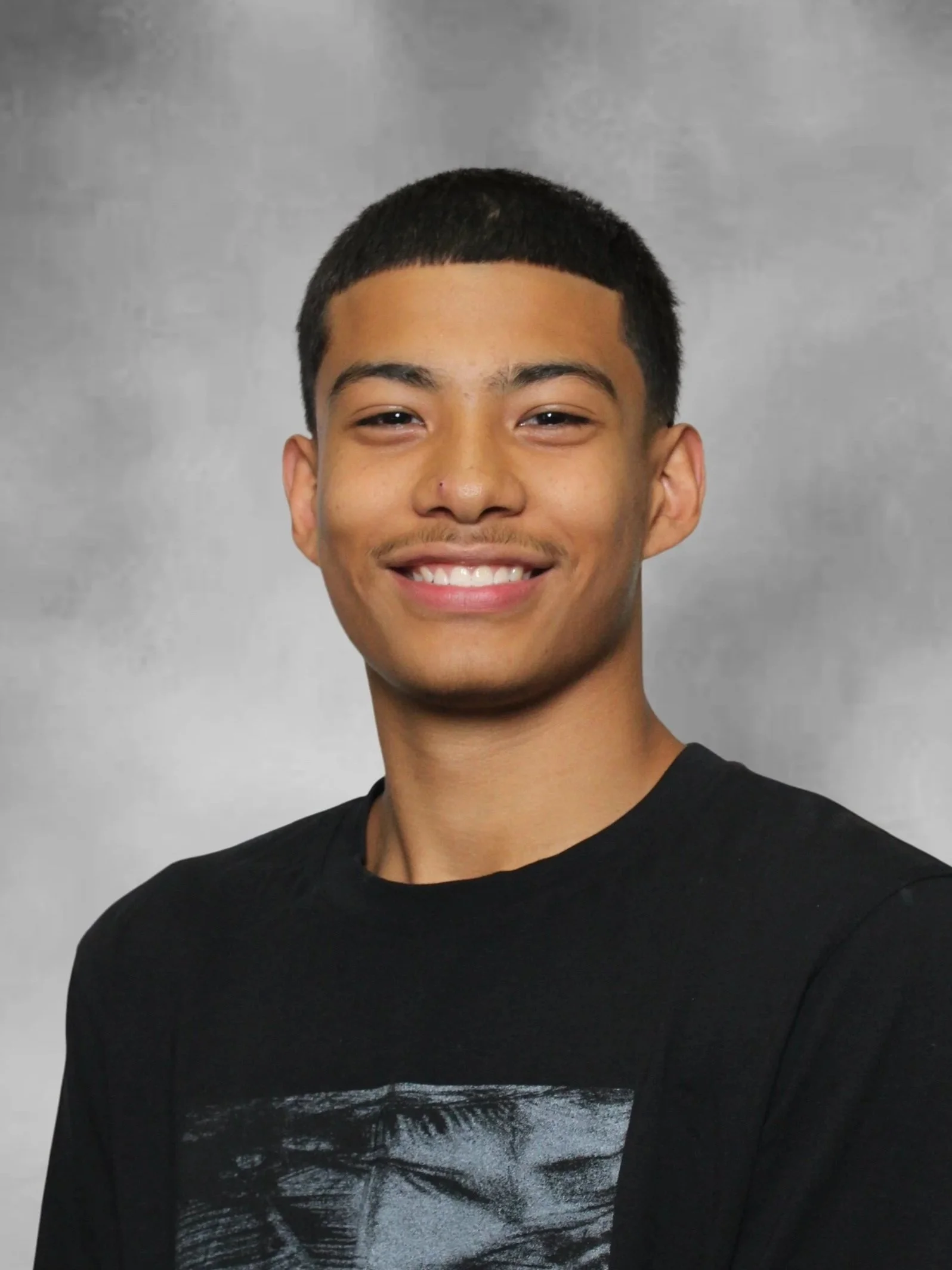 Close-up of a smiling young man with short black hair, wearing a black T-shirt with a graphic design, standing against a plain gray background.