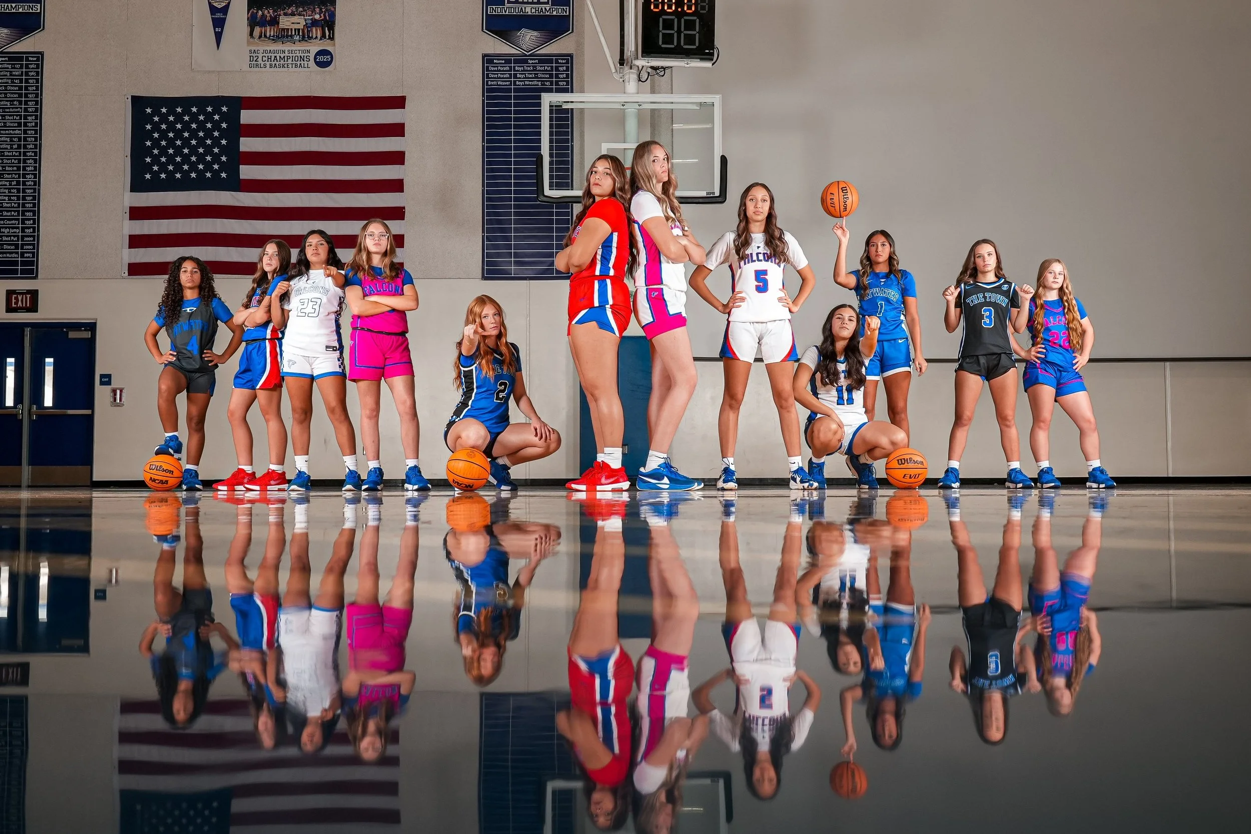 Group of young female basketball players in colorful uniforms standing in a gymnasium with a basketball court, USA flag, and banners. Some players hold basketballs, posing confidently with arms crossed or fists raised.
