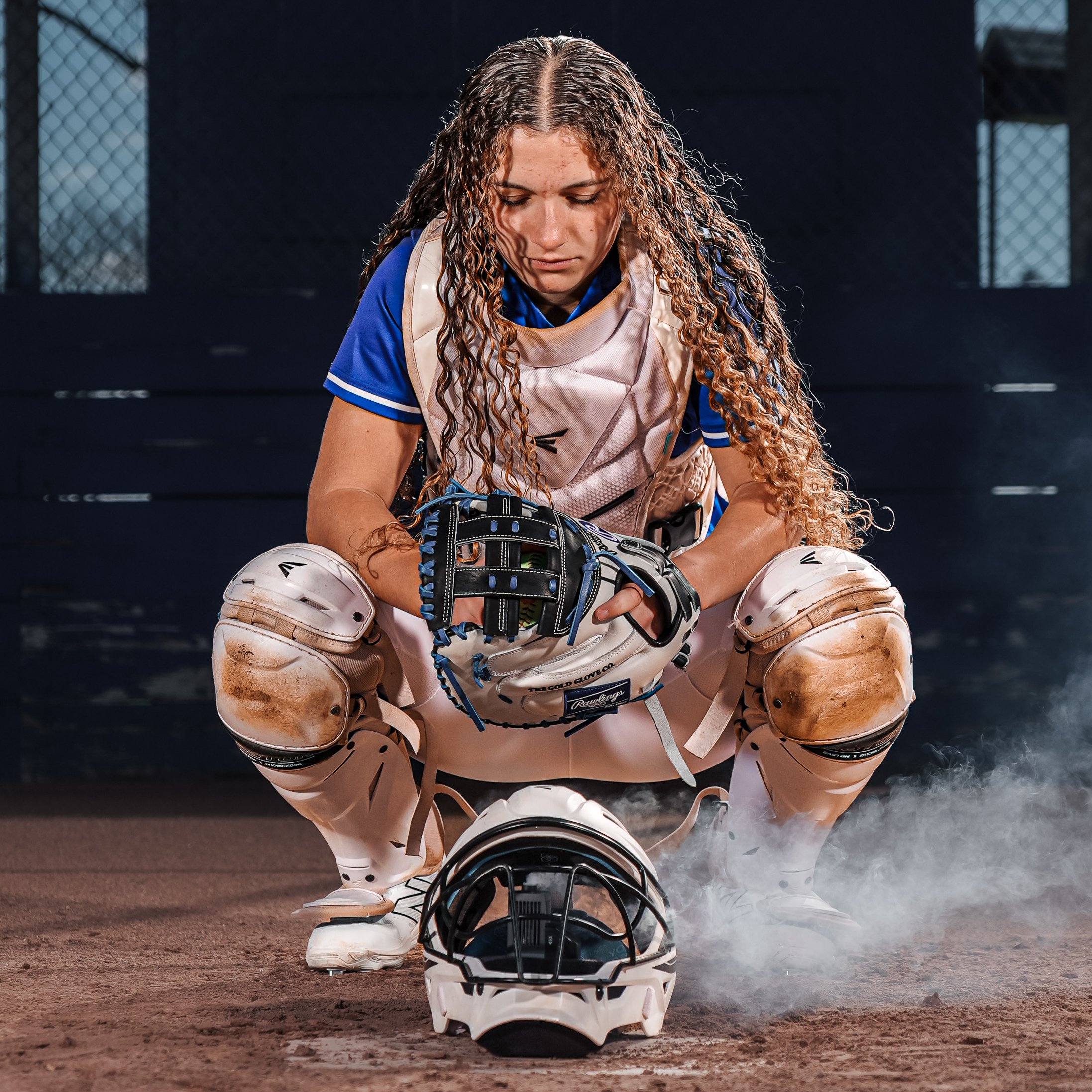 A female baseball catcher crouching on the field, wearing a pink catching gear and holding a black baseball glove, with a determined expression and head down, on a dirt baseball field with a chain-link fence in the background.