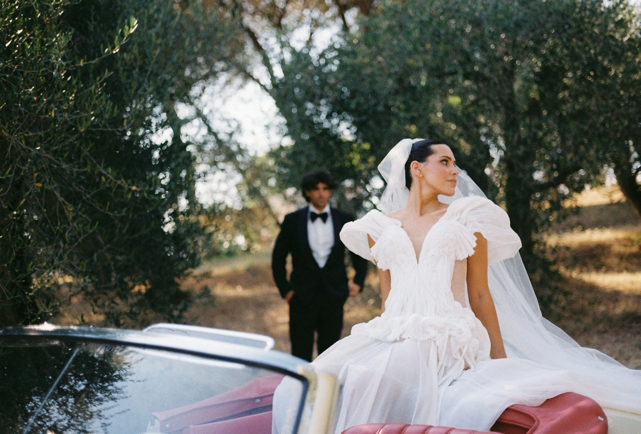 editorial film portrait of bride sitting in vintage car with groom in the background