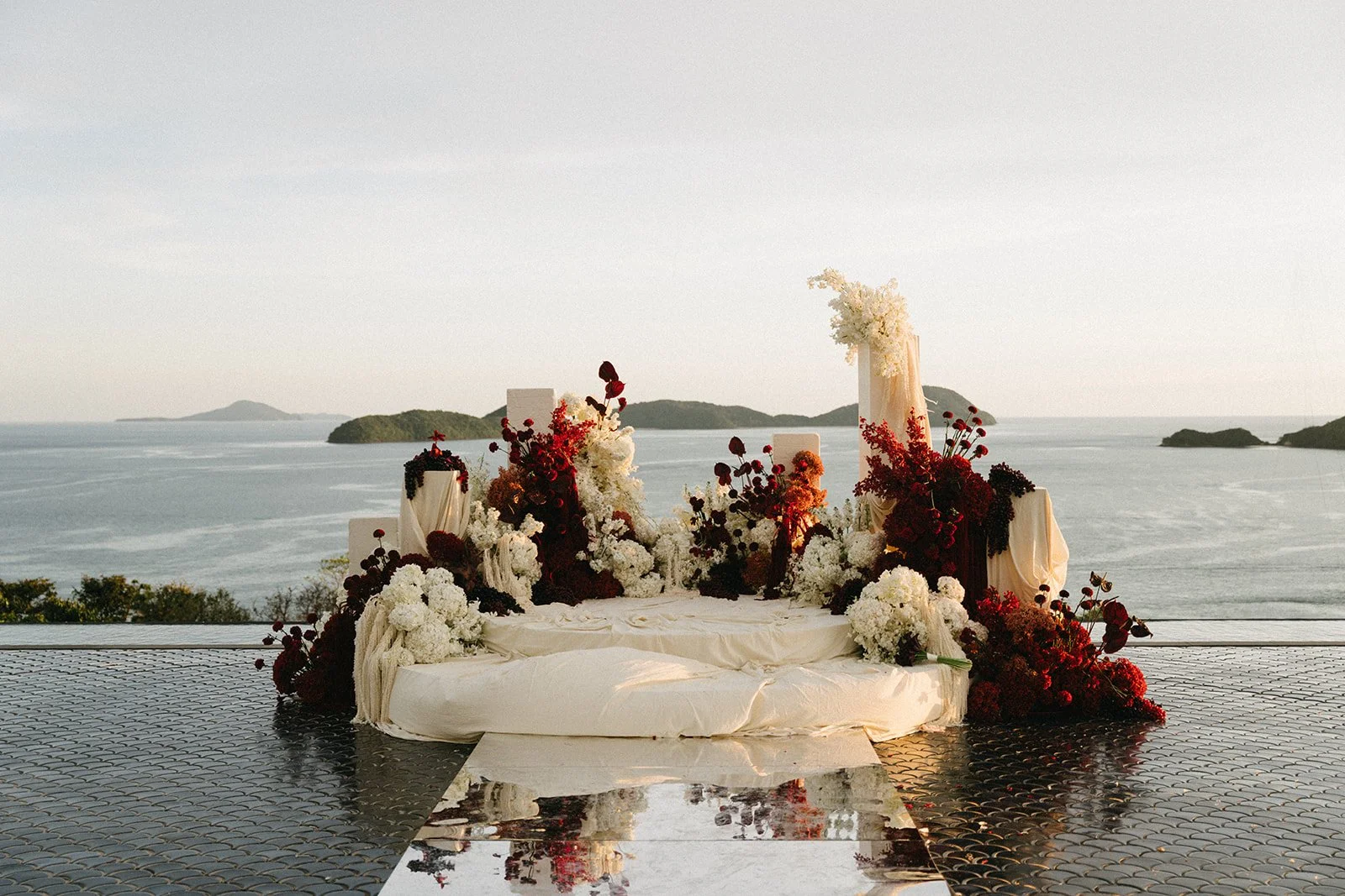 Infinity pool wedding ceremony setup at Sri Panwa with red, plum, and ivory floral installation overlooking the Andaman Sea