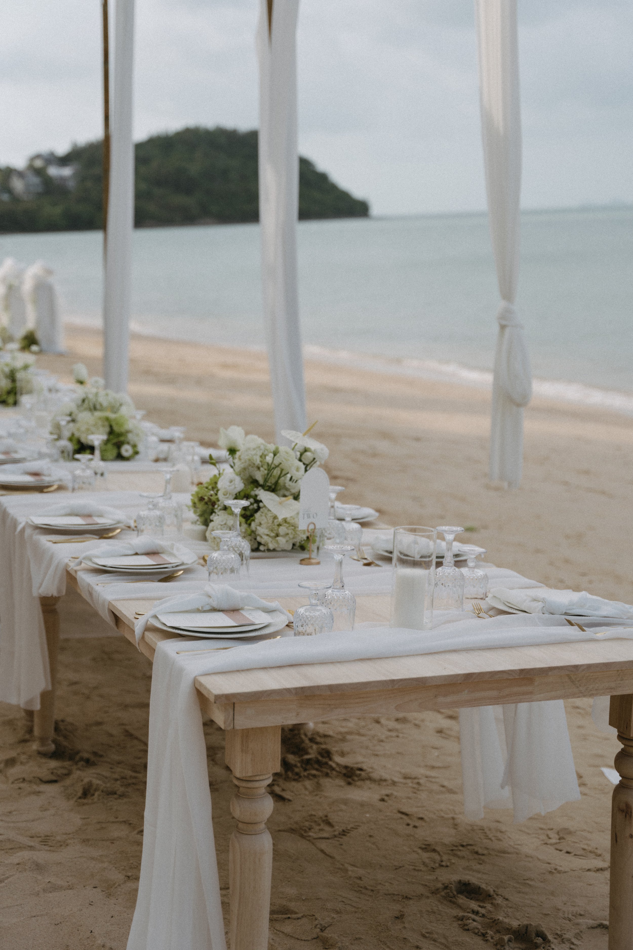 Long white banquet table with wooden chairs and floral arrangements on Bangrak Beach overlooking the ocean, Koh Samui