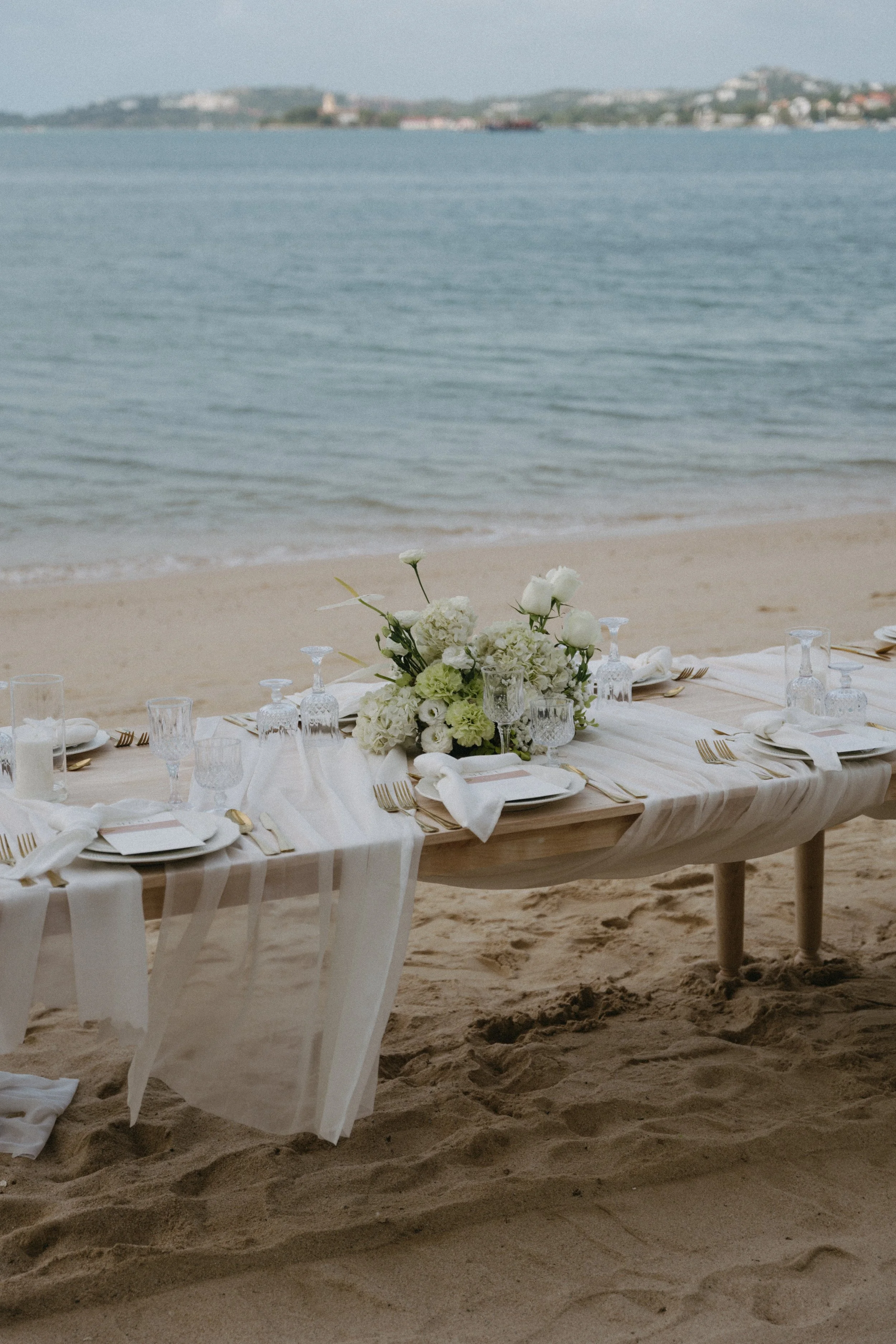 White linen dining table with green and white floral centrepiece set on the sand at Bangrak Beach destination wedding, Koh Samui