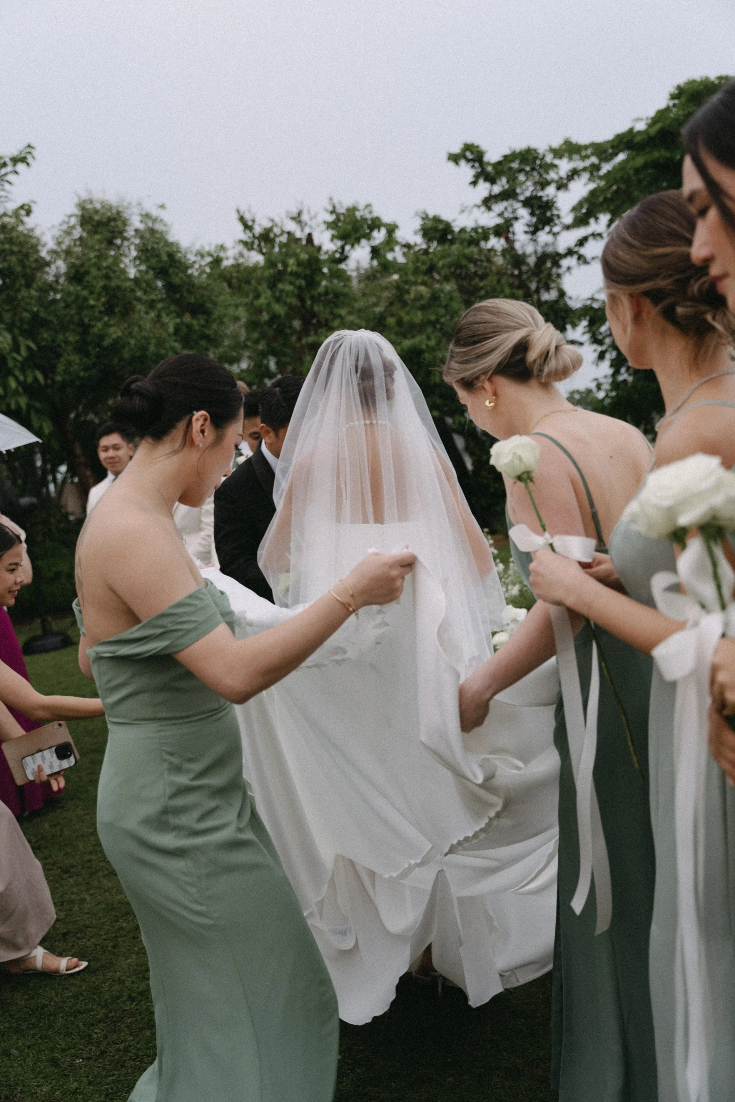Bridesmaids in sage green dresses adjusting bride's veil before rainy ceremony