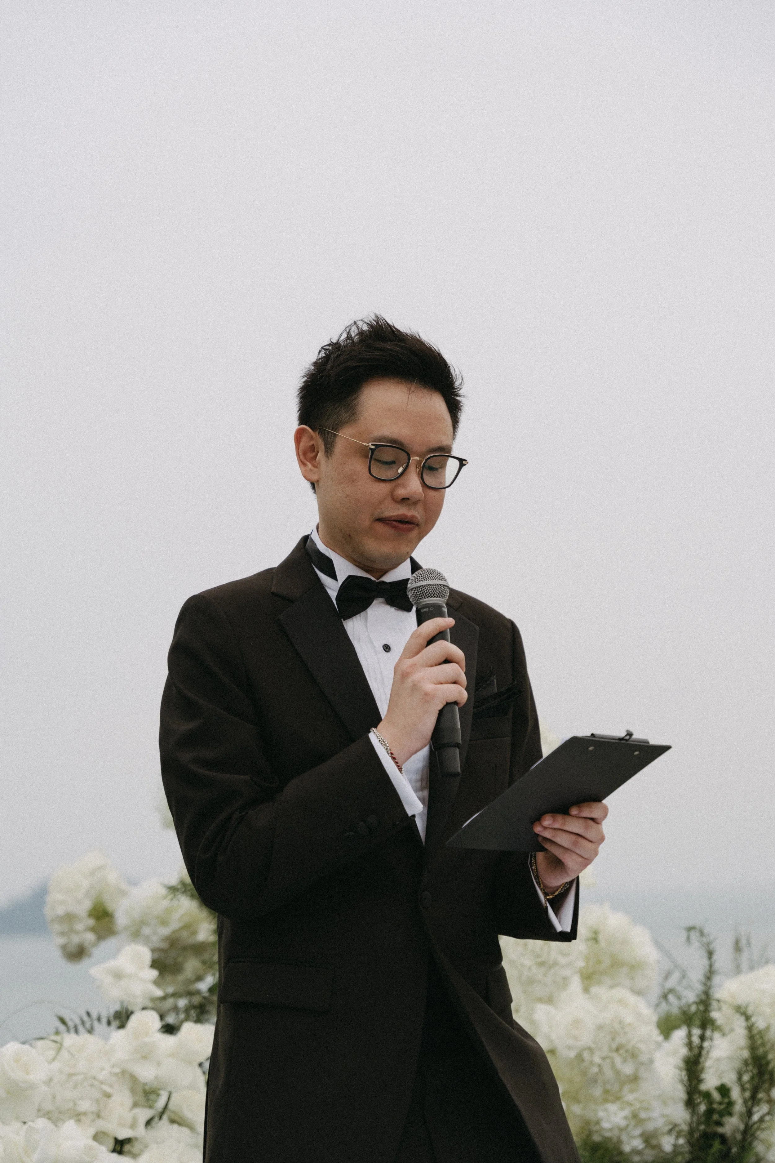 Celebrant in black tuxedo reading ceremony script, overcast Phuket wedding ceremony