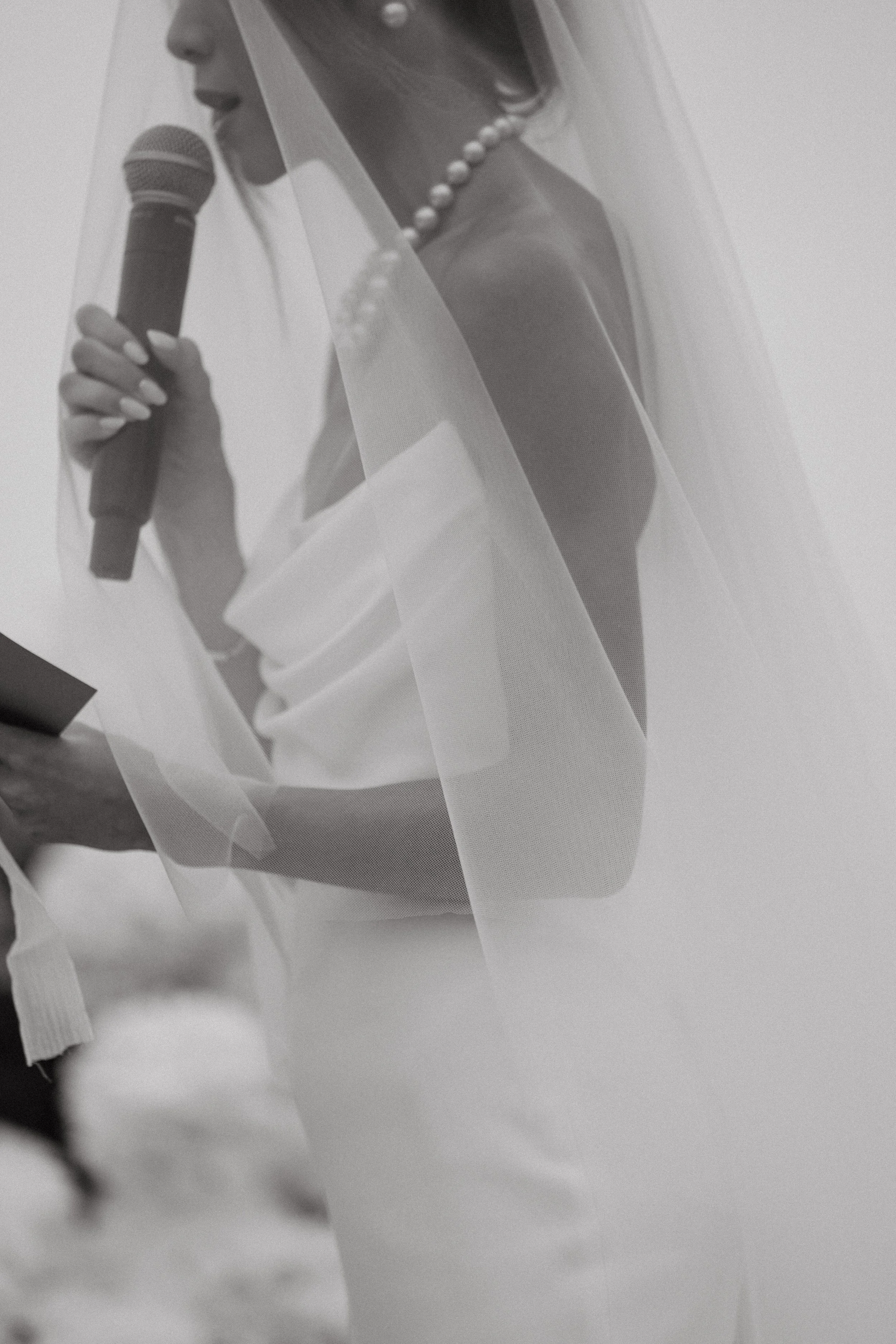 Close-up of bride holding mic under her veil during vows, black and white ceremony detail