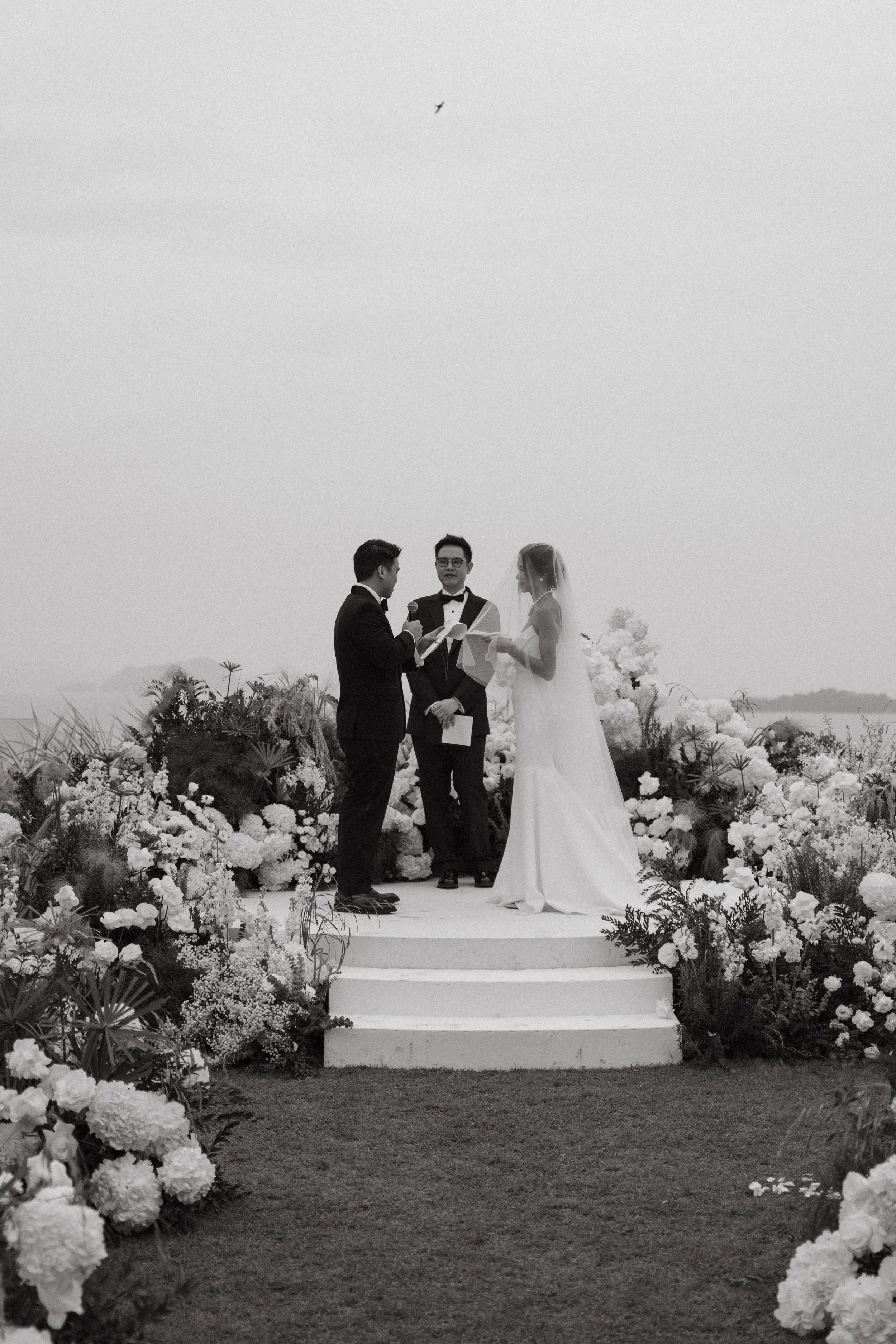 Bride and groom standing on round ceremony platform surrounded by white florals with phang nga bay backdrop
