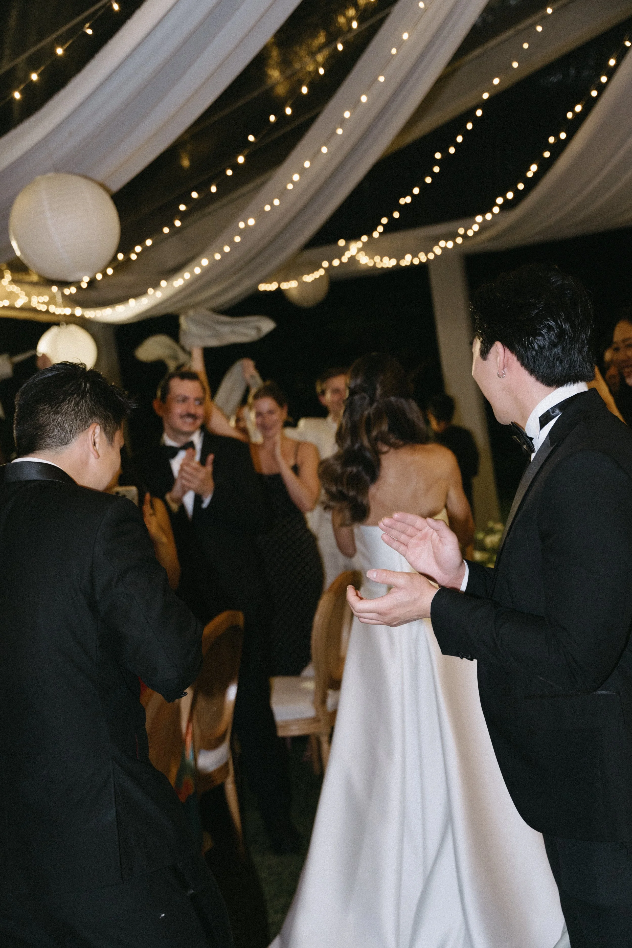 Wedding guests clapping and celebrating as newlyweds enter reception under lantern lights