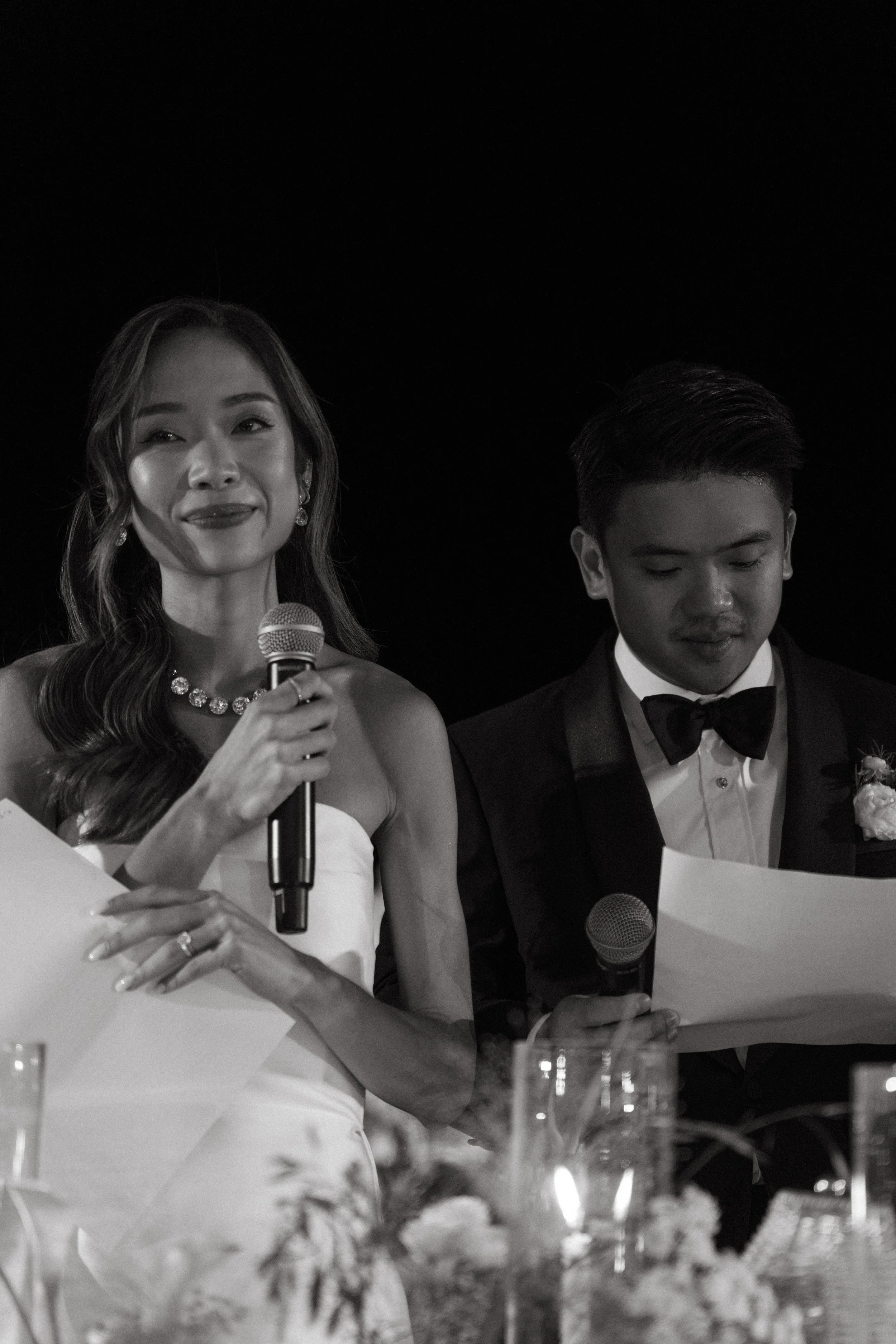  Bride and groom standing at reception table making wedding speech, microphone in hand