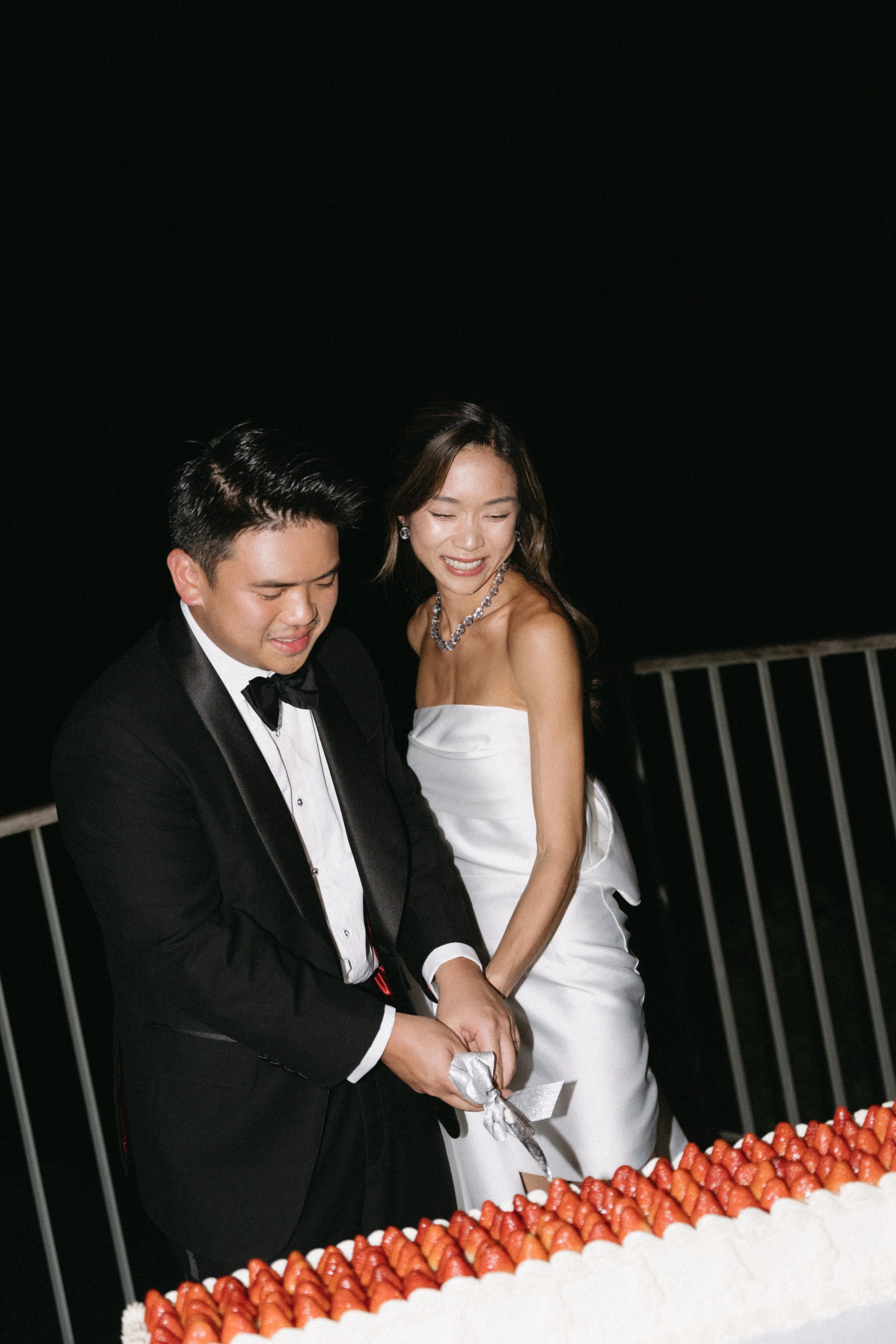 Bride and groom cutting strawberry-topped wedding cake together at night reception