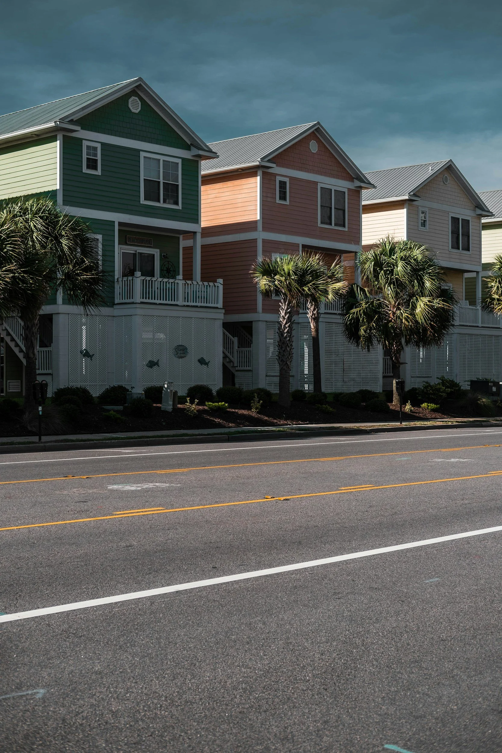 Colorful beach houses in pastel shades of green, pink, and beige with palm trees in front, along a road.