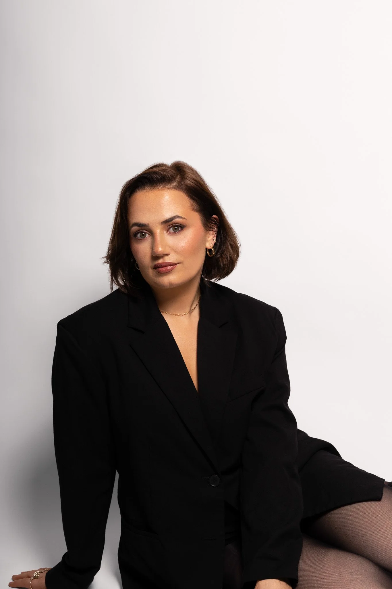 Annalee Delaney Photographer is sitting down in a photogrpahy studio against a white background wearing a black blazer and looking at the camera