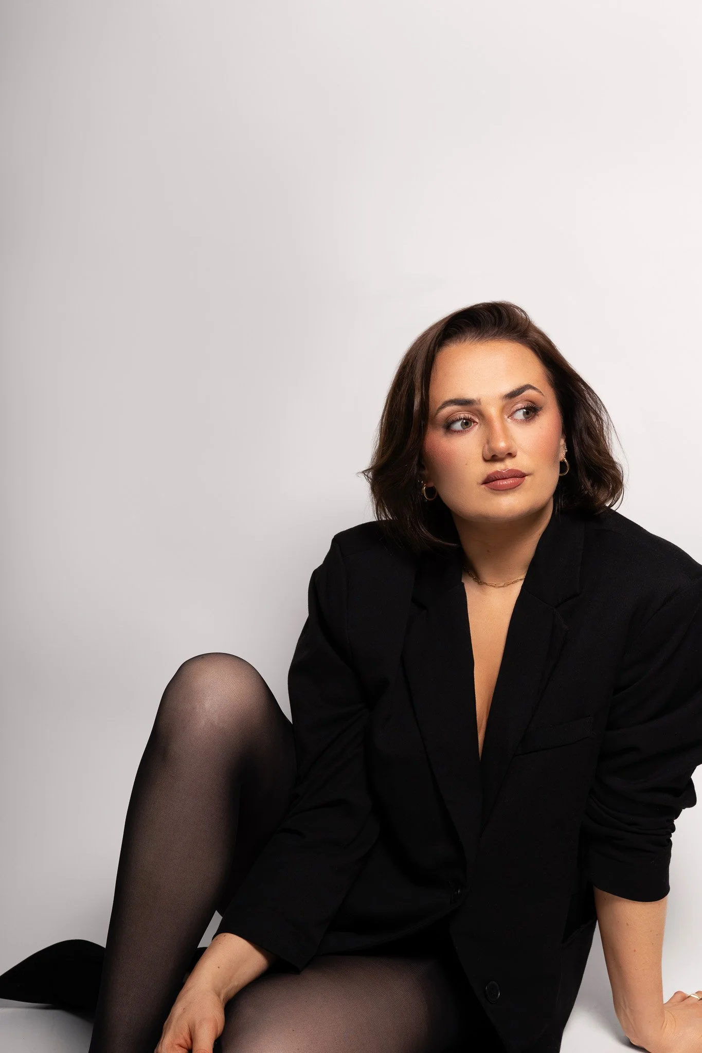 Annalee Delaney the photographer  is sitting down in a photography studio against a white backdrop wearing a black blazer looking to the right, away from the camera