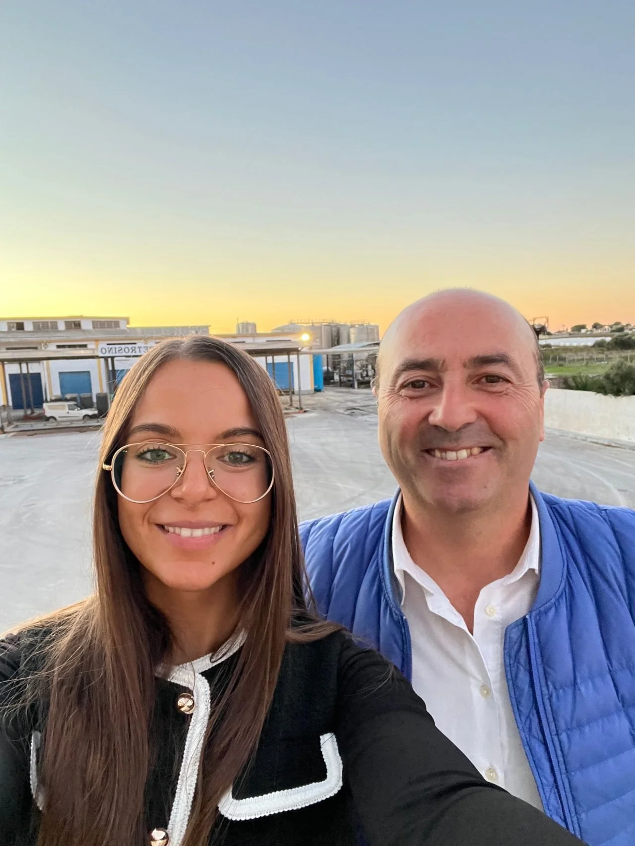 A selfie of a woman and a man smiling outdoors at sunset, with an industrial area in the background.