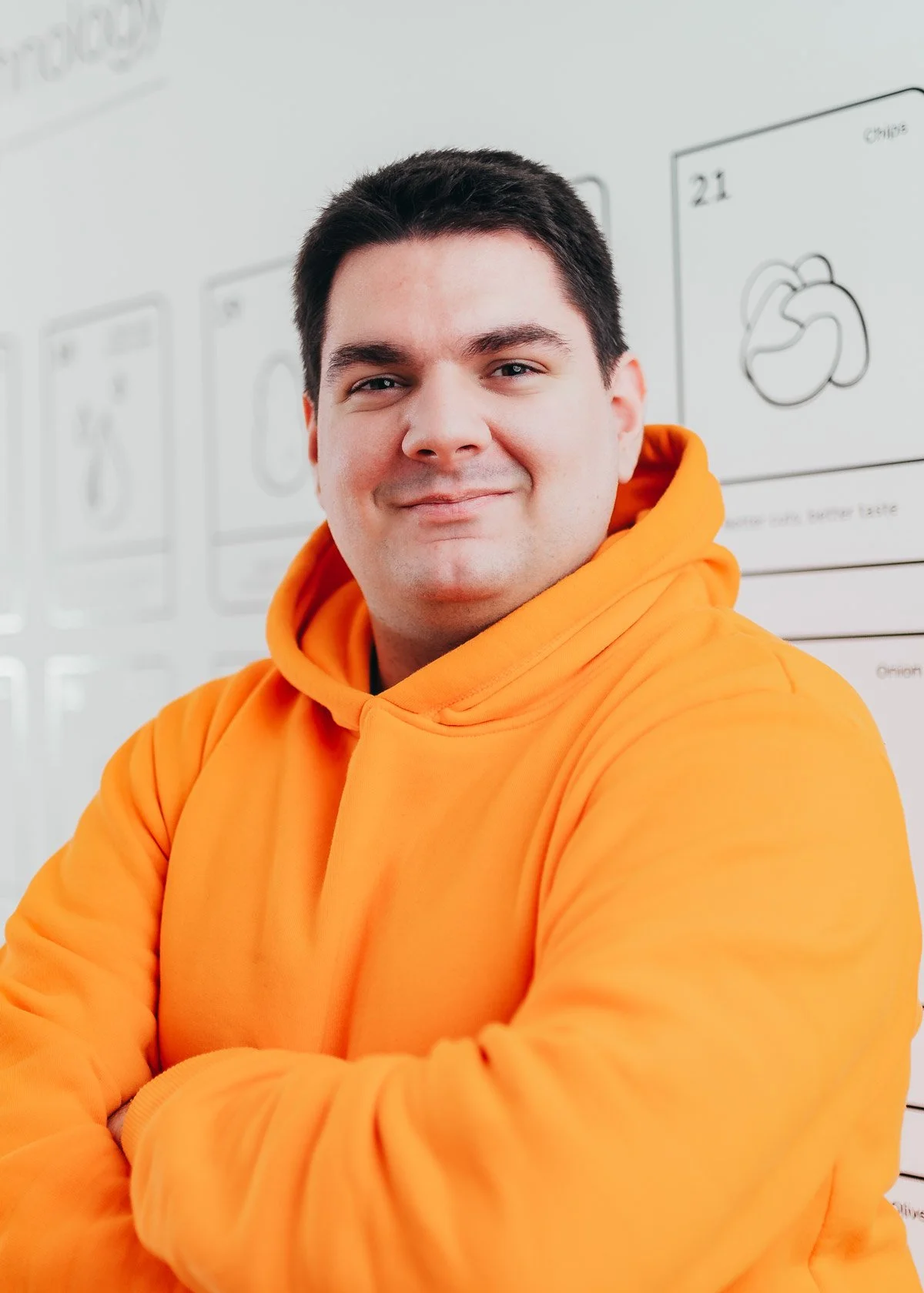 Man in orange hoodie smiling with arms crossed, standing in front of a whiteboard with drawings.