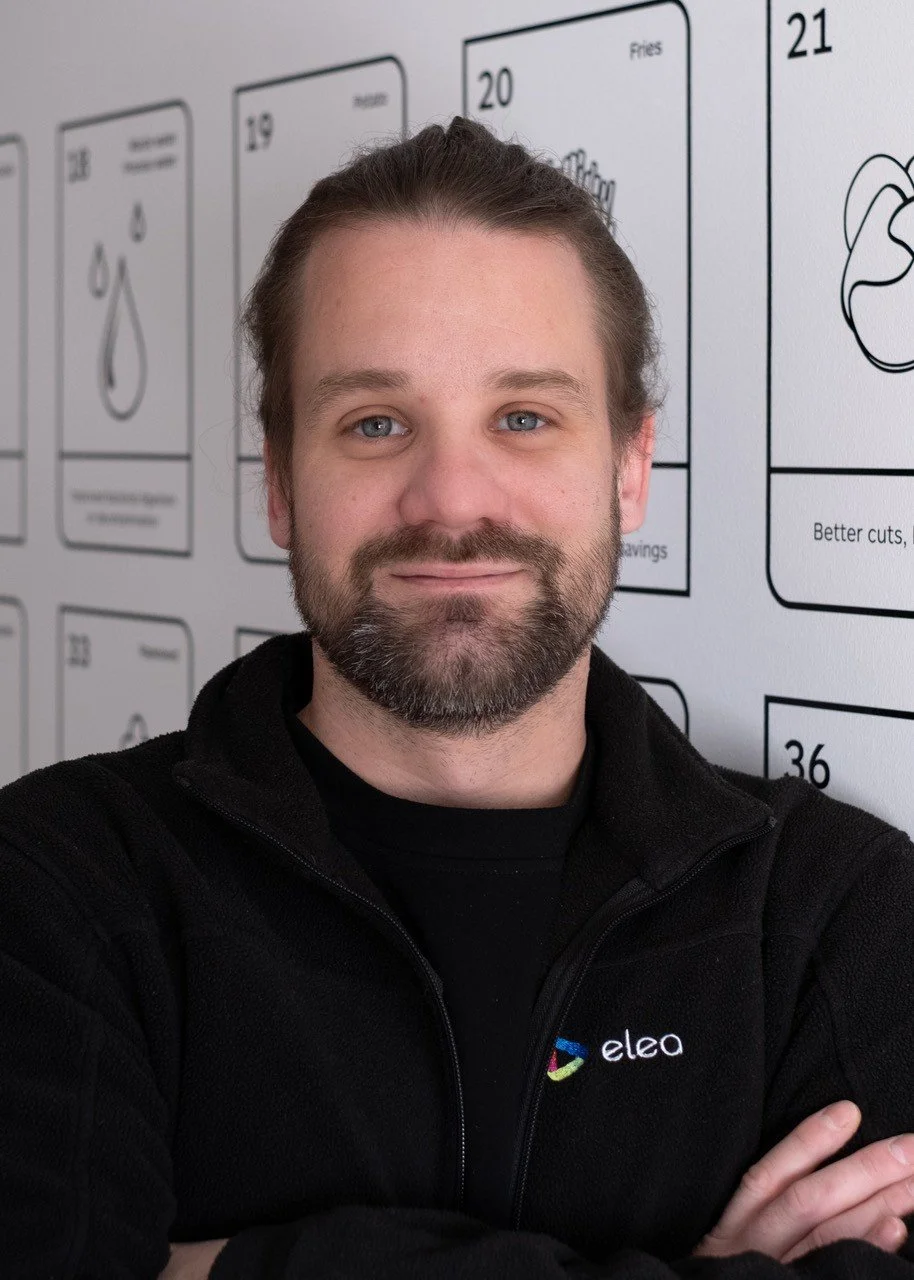 Person wearing a black zip-up jacket with "elea" logo, smiling in a bright indoor setting.