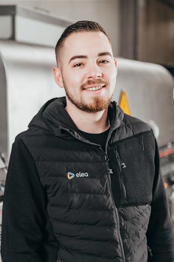 A young man with a beard and short hair smiling at the camera, wearing a black vest with an 'elea' logo, in an indoor industrial or warehouse setting.
