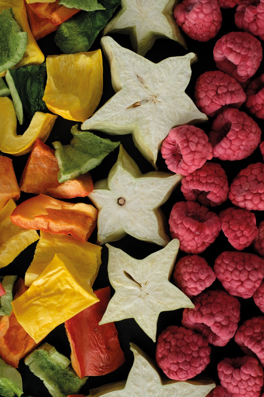 Close-up of dried starfruit slices, colorful dried bell peppers, and fresh raspberries arranged in a pattern on a dark background.