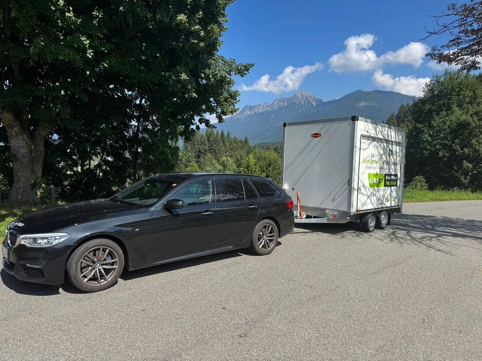 A black BMW station wagon parked on the side of a road with a trailer attached. The trailer has a white exterior with green and black text promoting electric vehicle charging. The background features a mountain range, trees, and a partly cloudy sky.