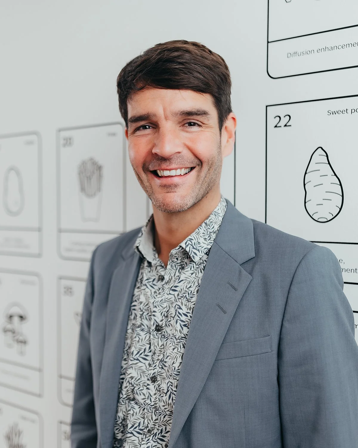 Person in a gray suit with a patterned shirt standing in front of a wall with illustrations and text, smiling at the camera.