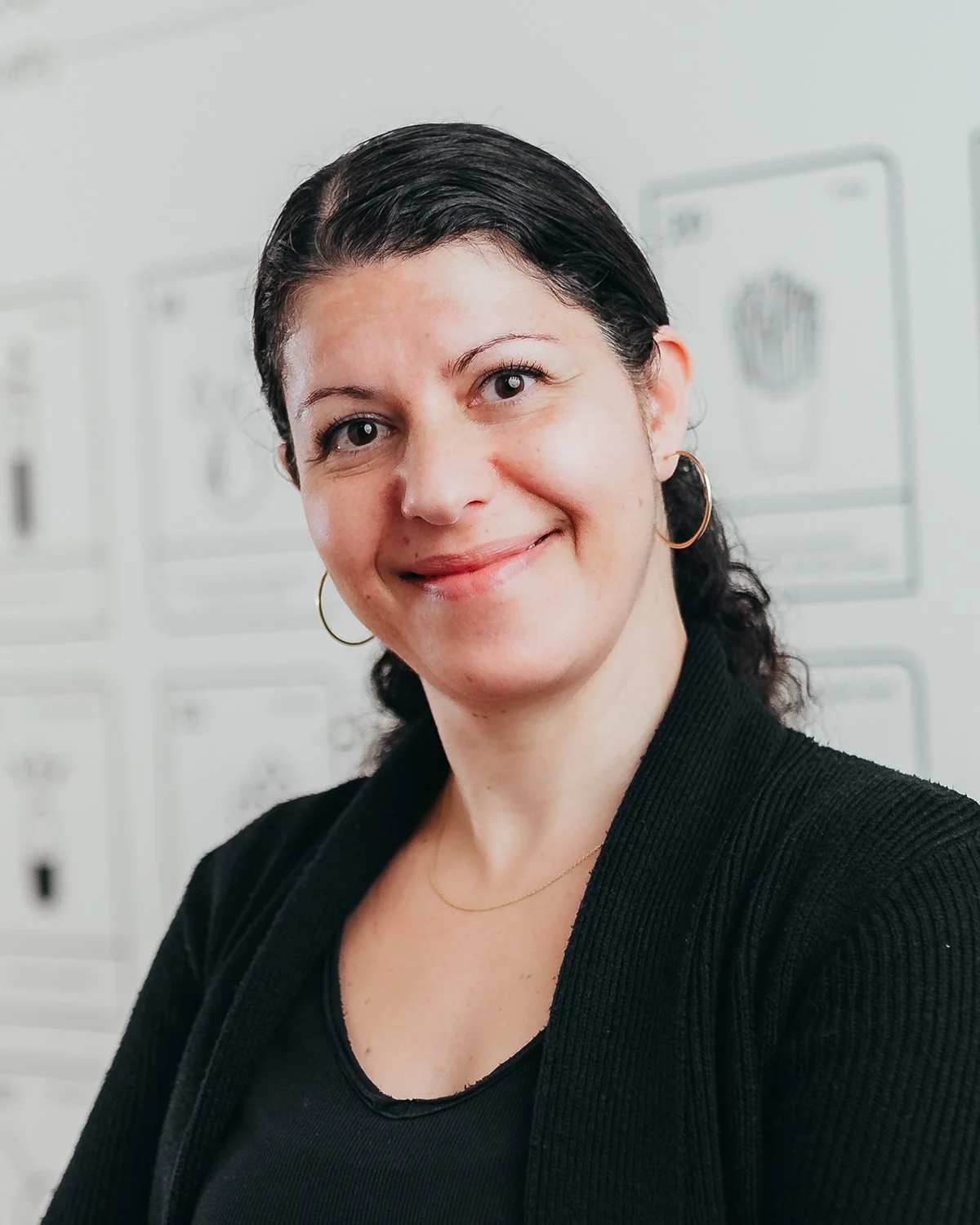 Smiling person with dark hair in a black top and hoop earrings, standing in front of a blurred background with charts or diagrams.