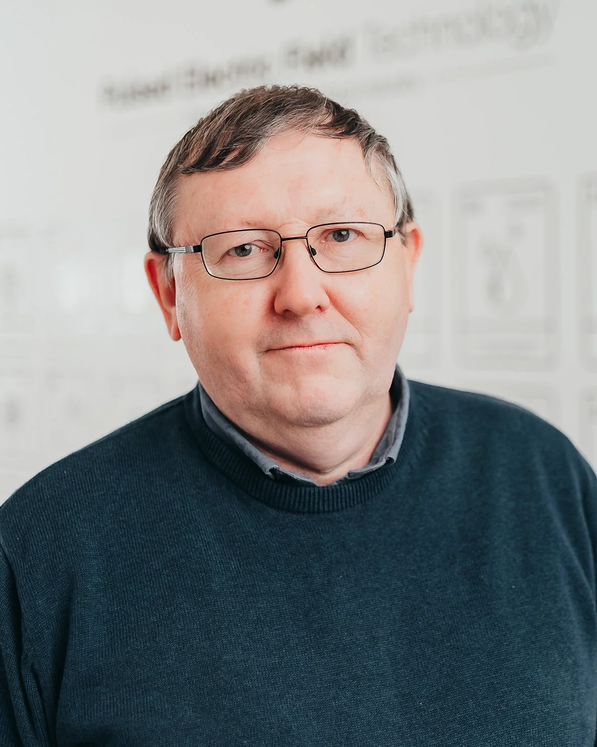 Middle-aged man with short hair and glasses wearing a dark sweater, standing against a blurred white background with text.