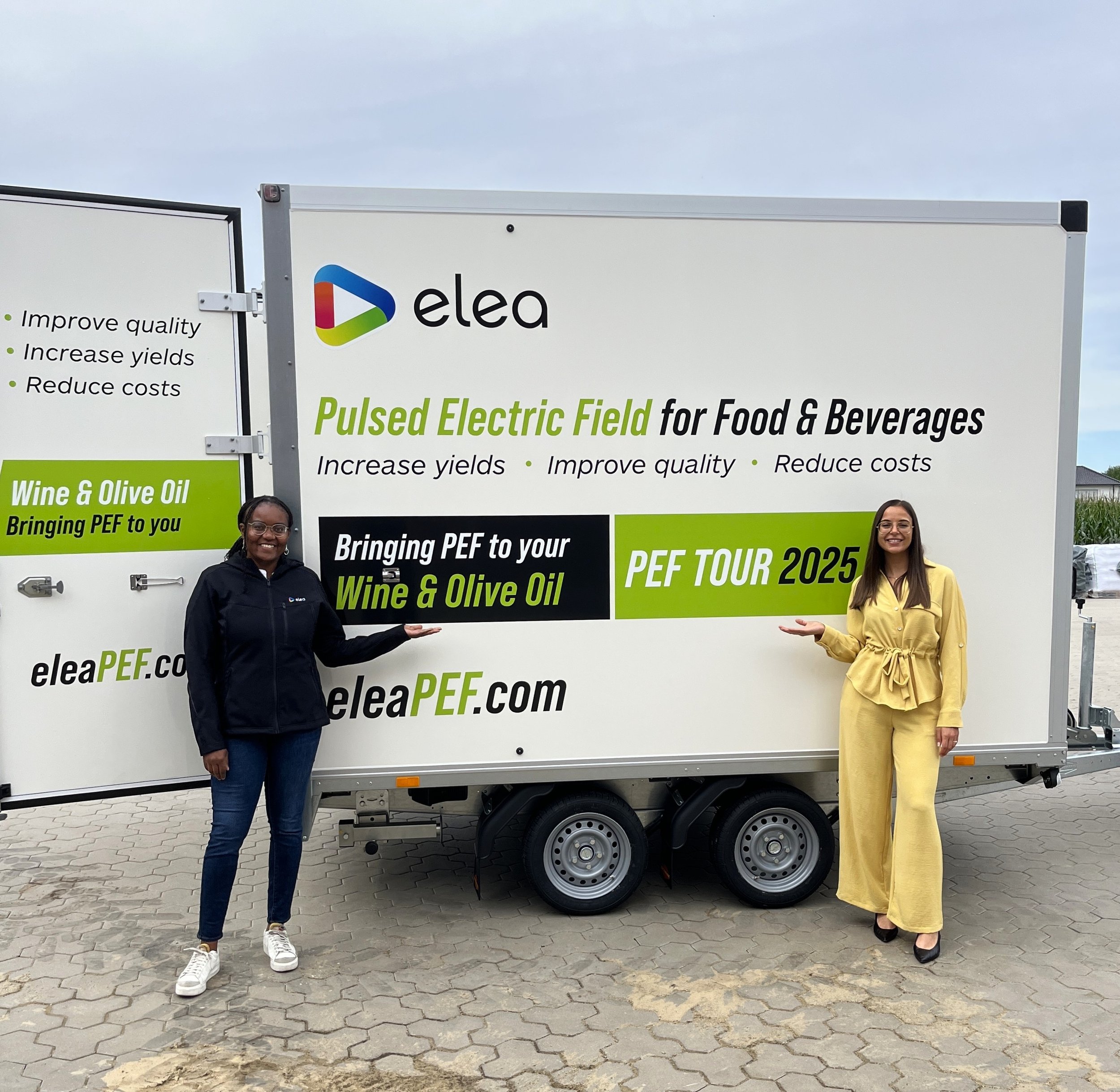 Four men standing in a factory or industrial setting engaged in conversation. One man in a yellow shirt is looking at a paper or object in his hands.