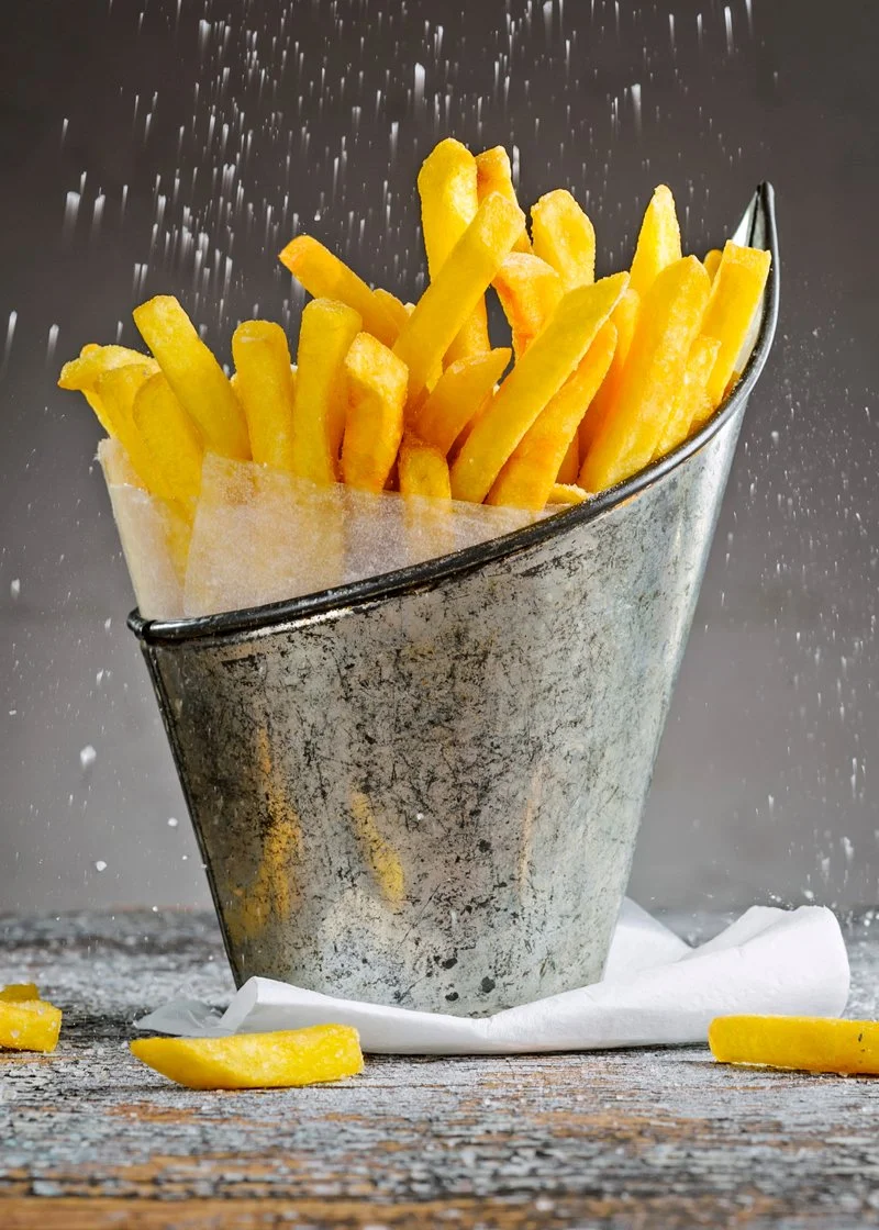 Golden French fries in a rustic metal container, dusted with salt, on a wooden surface.