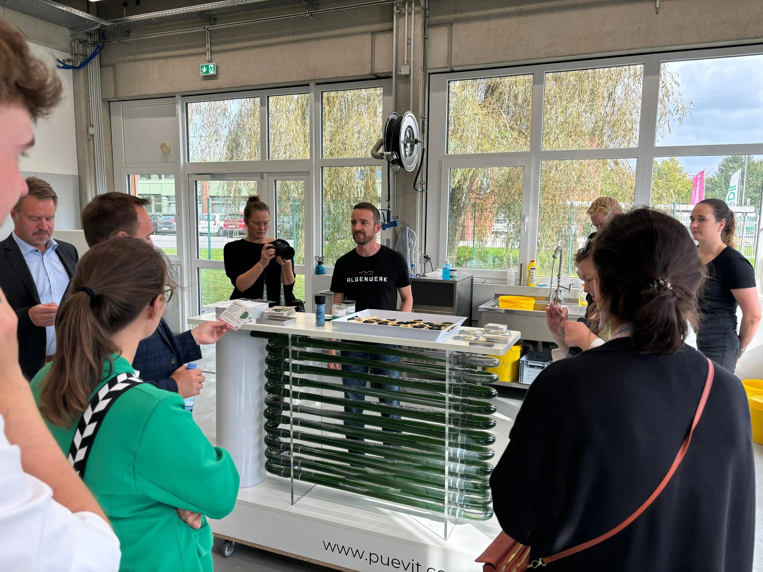 Group of people gathered around a science demonstration in a laboratory with large windows and green outdoor scenery. One person in the center appears to be an instructor explaining something, with a tray of small containers on the table.