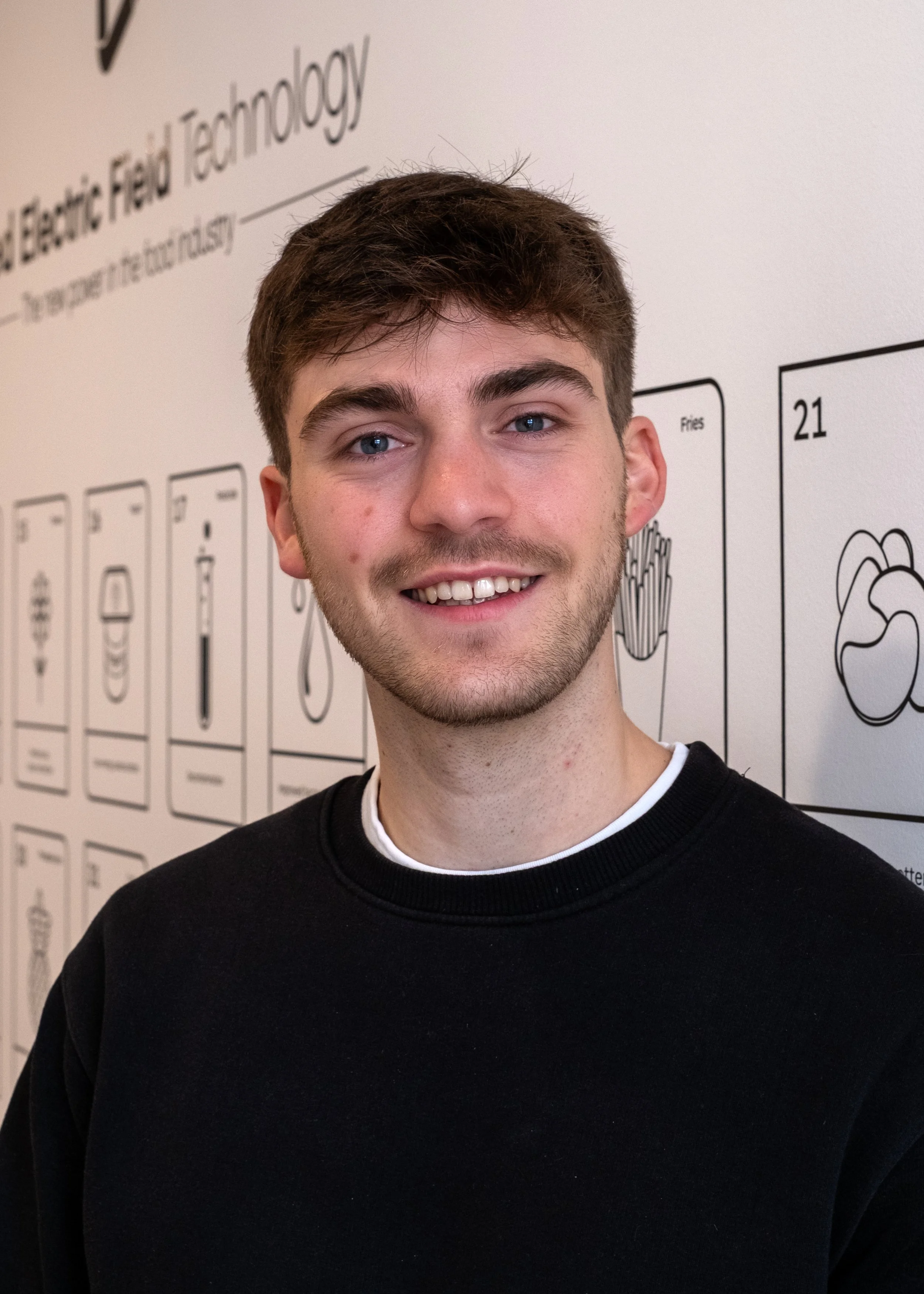 A young man with brown hair, blue eyes, and light skin smiling, standing in front of a white wall with illustrations and text about electric field technology.