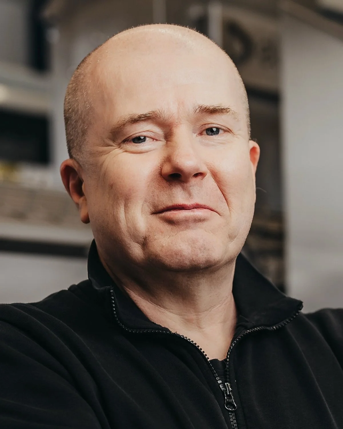 Bald man wearing a black jacket with a neutral expression, blurred background.