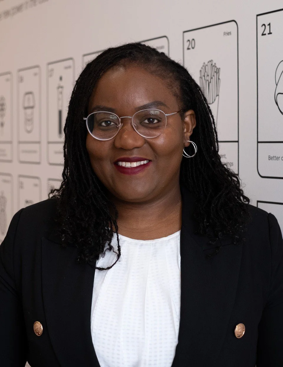 A person wearing glasses, a white shirt, and a black blazer with braided hair, smiling at the camera. The background appears to be a wall with graphics and text.